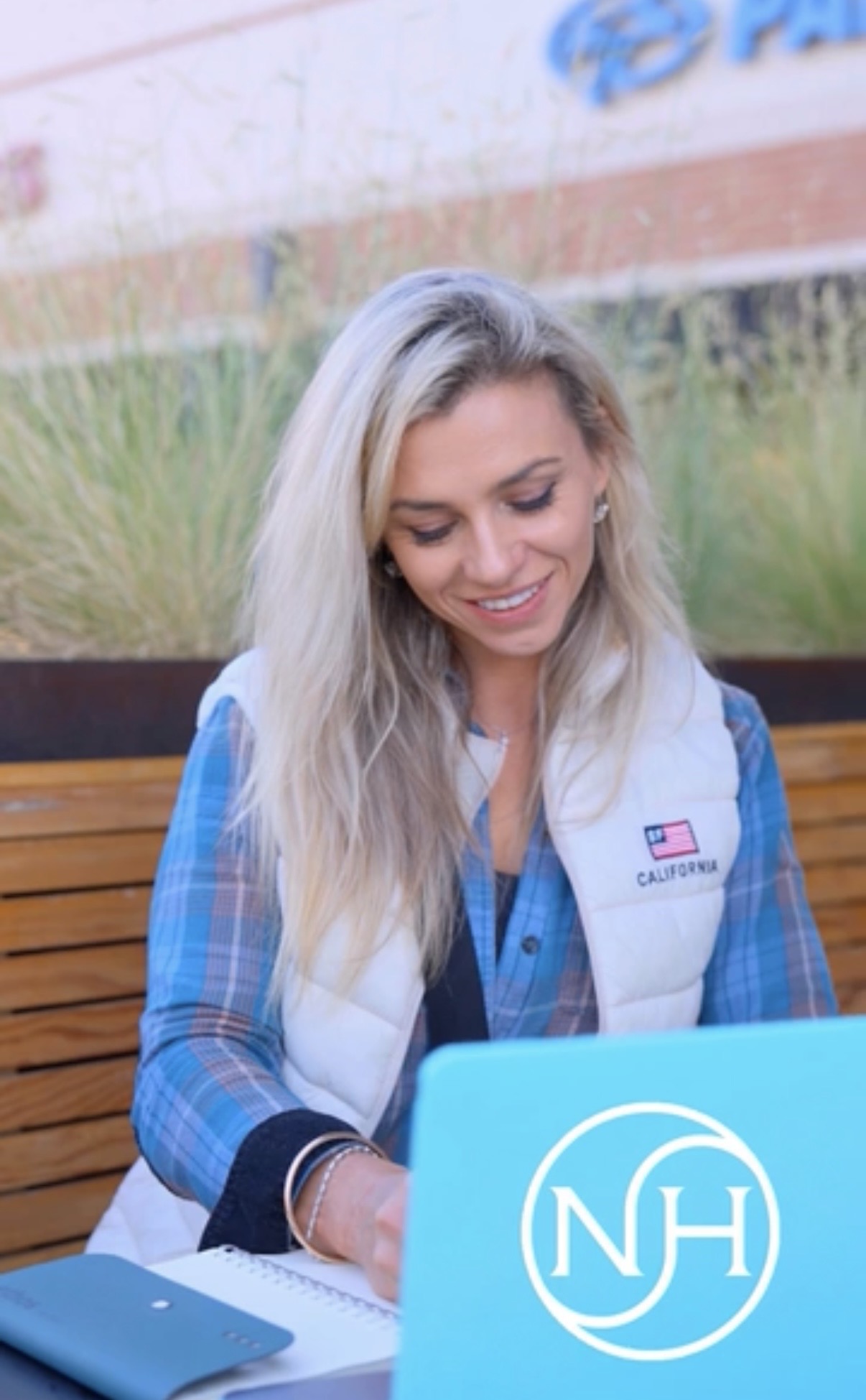 Young woman with long blonde hair smiling, sitting outdoors with a blue notebook and NH sign, wearing a vest and plaid shirt.