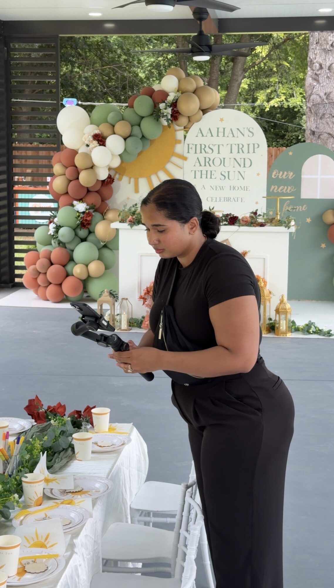 Woman in black outfit holding a tablet at decorated event table with floral arrangement and balloons, outdoor setting