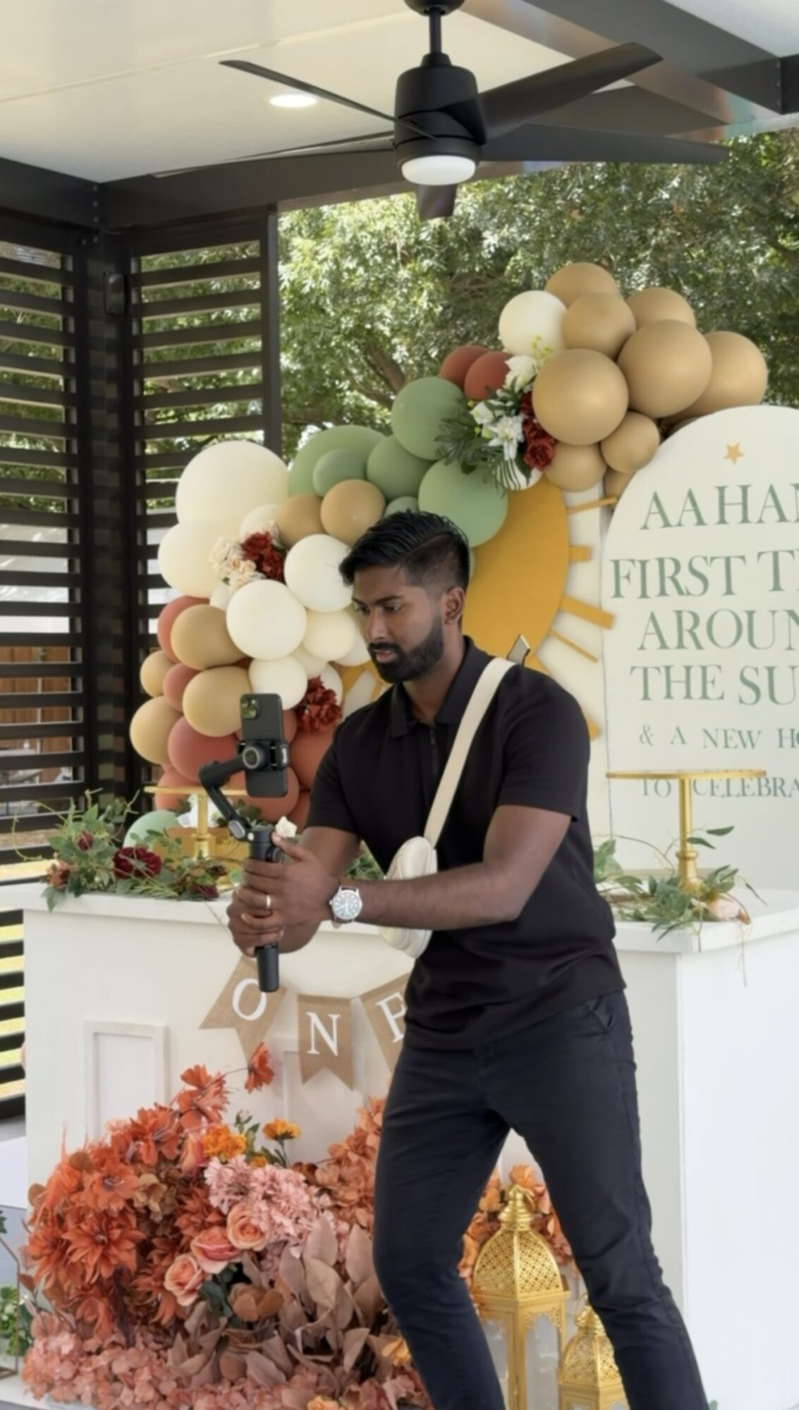 Man standing outdoors near decorated table with balloons and flowers, holding a drink, trees visible in background.
