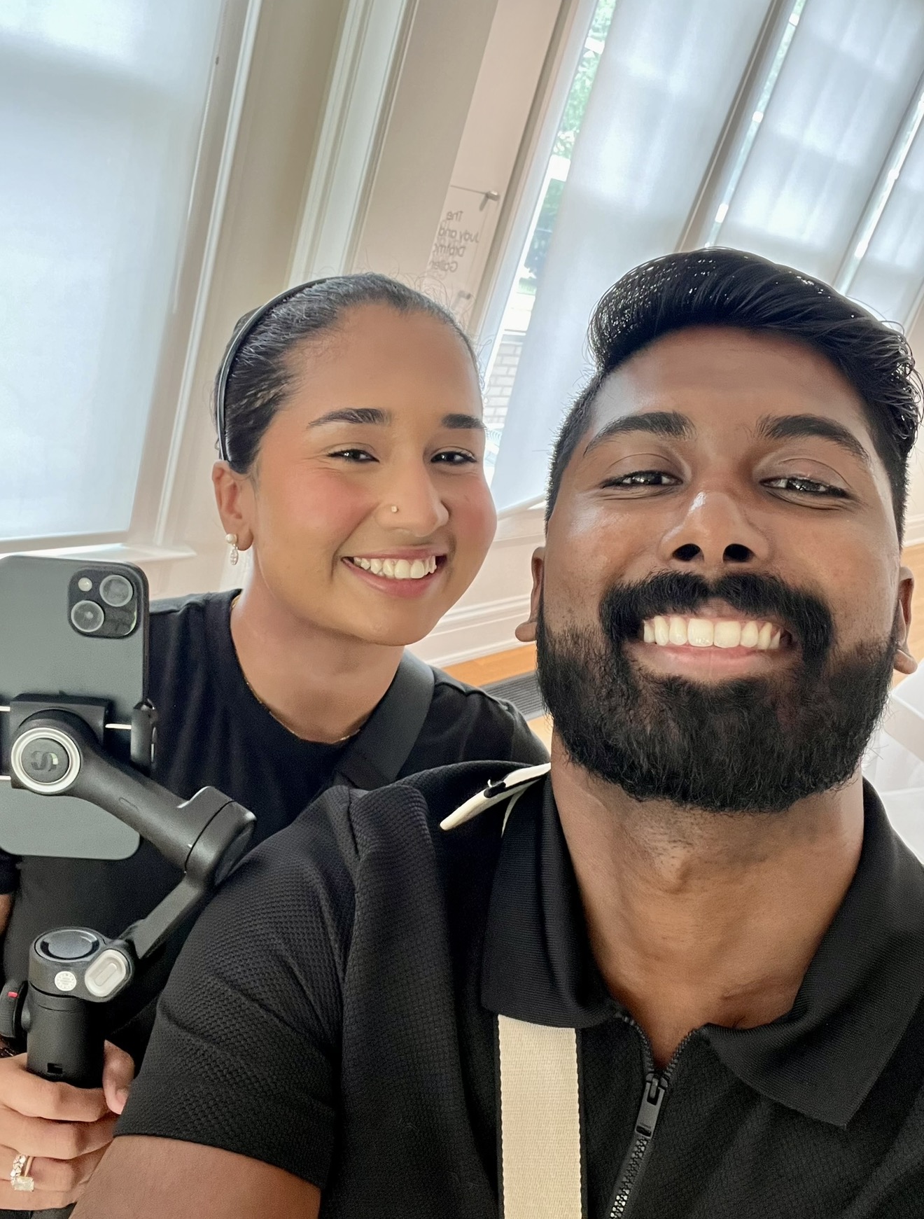 Two people smiling for a selfie indoors, with a woman holding a camera or phone on a stabilizer, and a man in front.