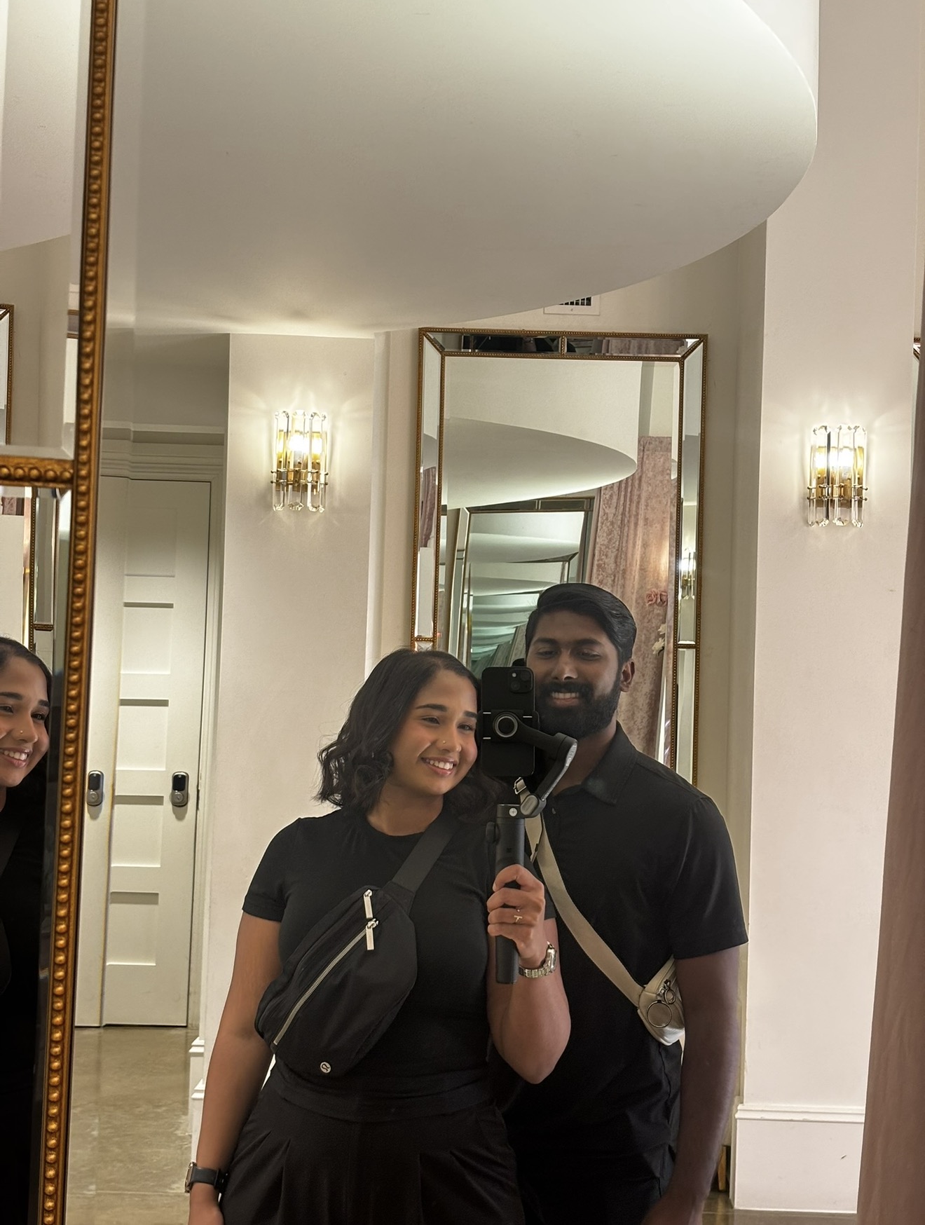 Two people taking a mirror selfie in a well-lit room with elegant decor, including wall sconces and mirrors.
