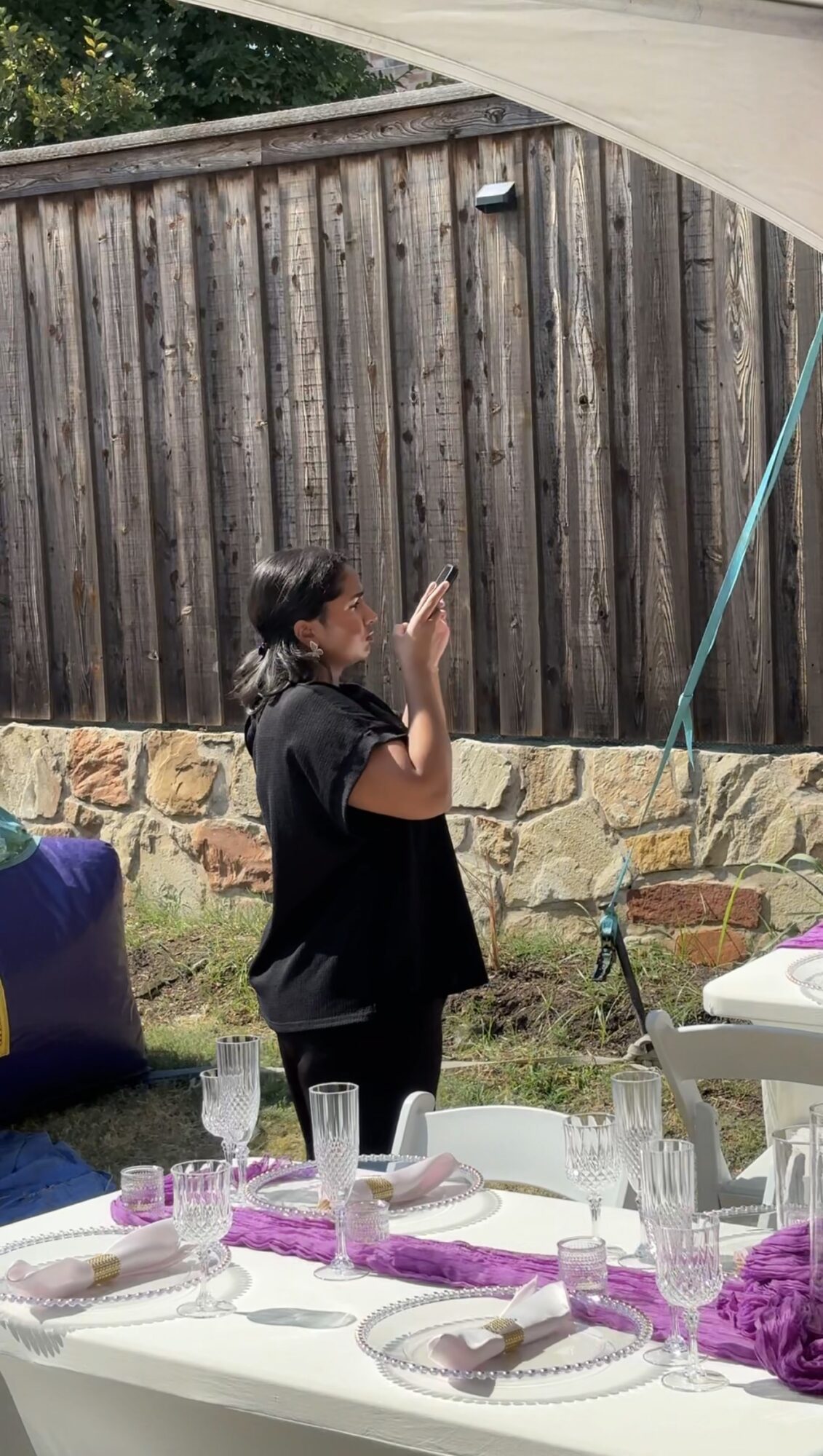 Person standing outdoors near a wooden fence, with a table set with glasses and purple decorations in the foreground.