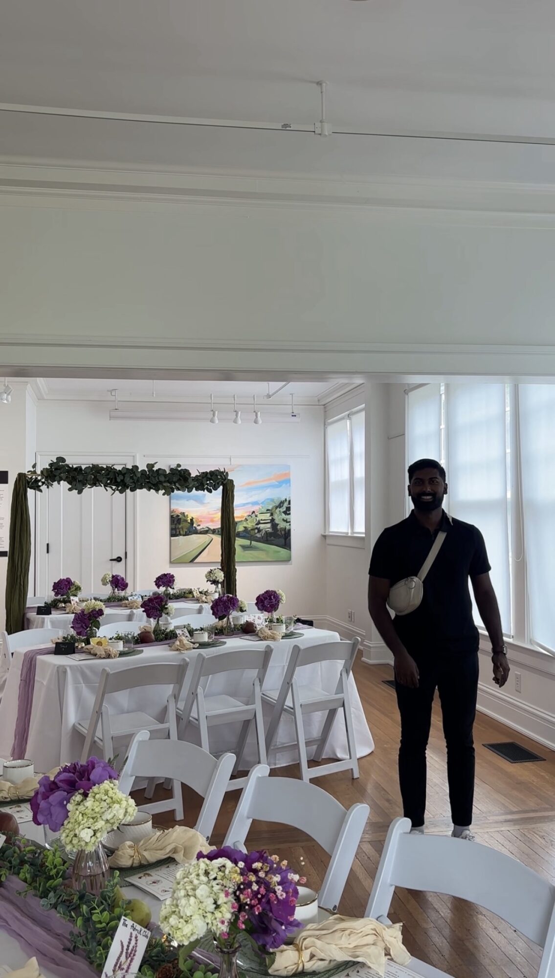Person standing near decorated table with flowers and chairs in a bright room with large windows and landscape painting.