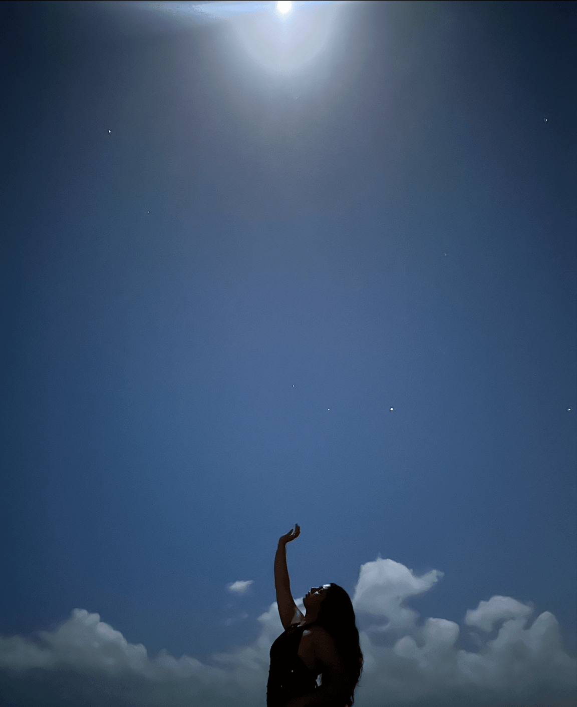 Person standing outdoors under a bright moonlit sky with clouds, raising an arm upward.