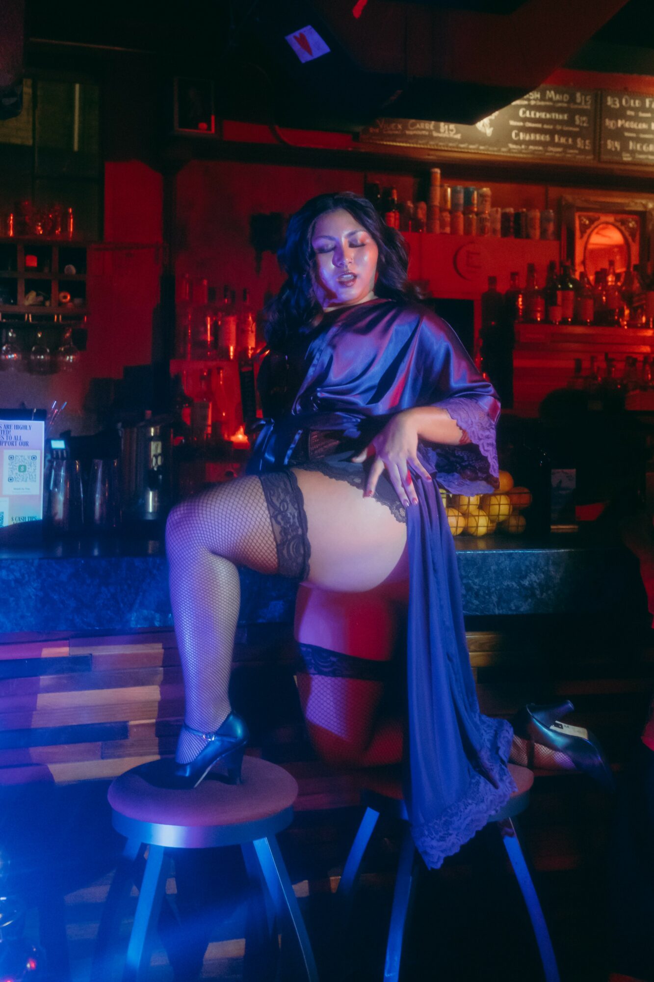 Woman in a dark blue satin robe and lace stockings poses on a bar counter in a dimly lit bar, with red lighting.