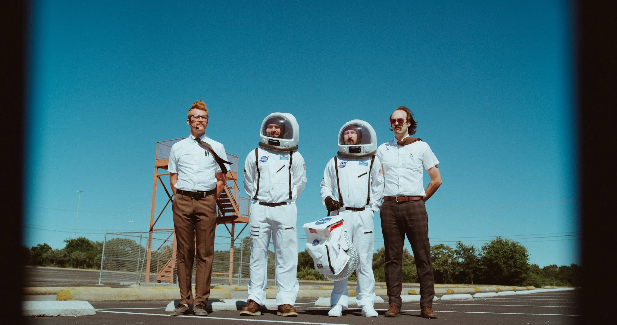 Three people in astronaut suits and two men in casual clothing stand outdoors against a blue sky.