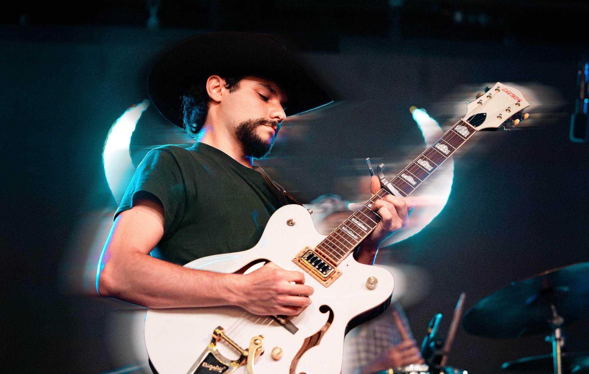 Person playing an electric guitar on stage with colorful lighting, wearing a hat and dark shirt.