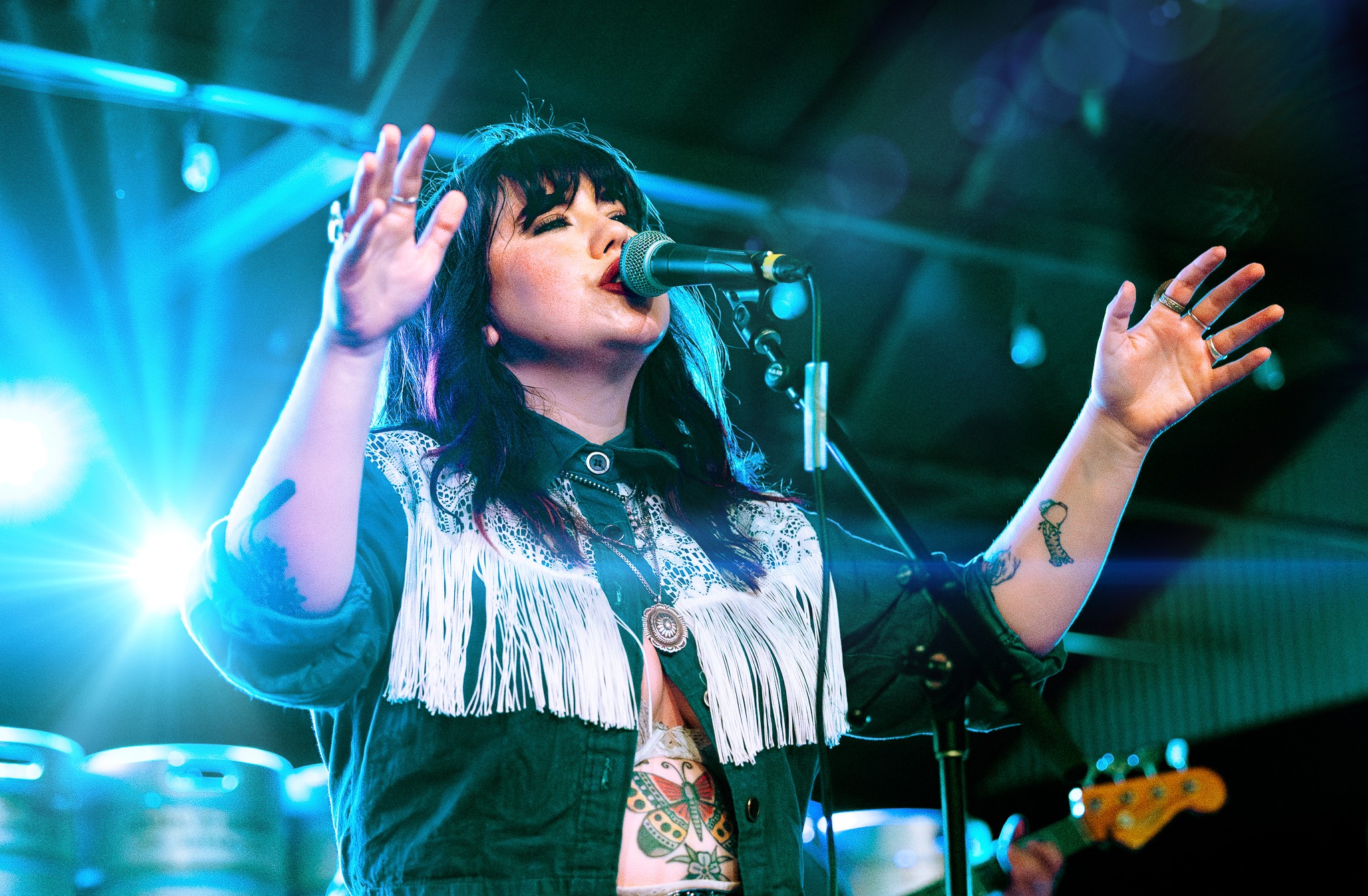 Female singer with black hair and fringe performs on stage with arms raised, microphone in front, bright stage lights behind.