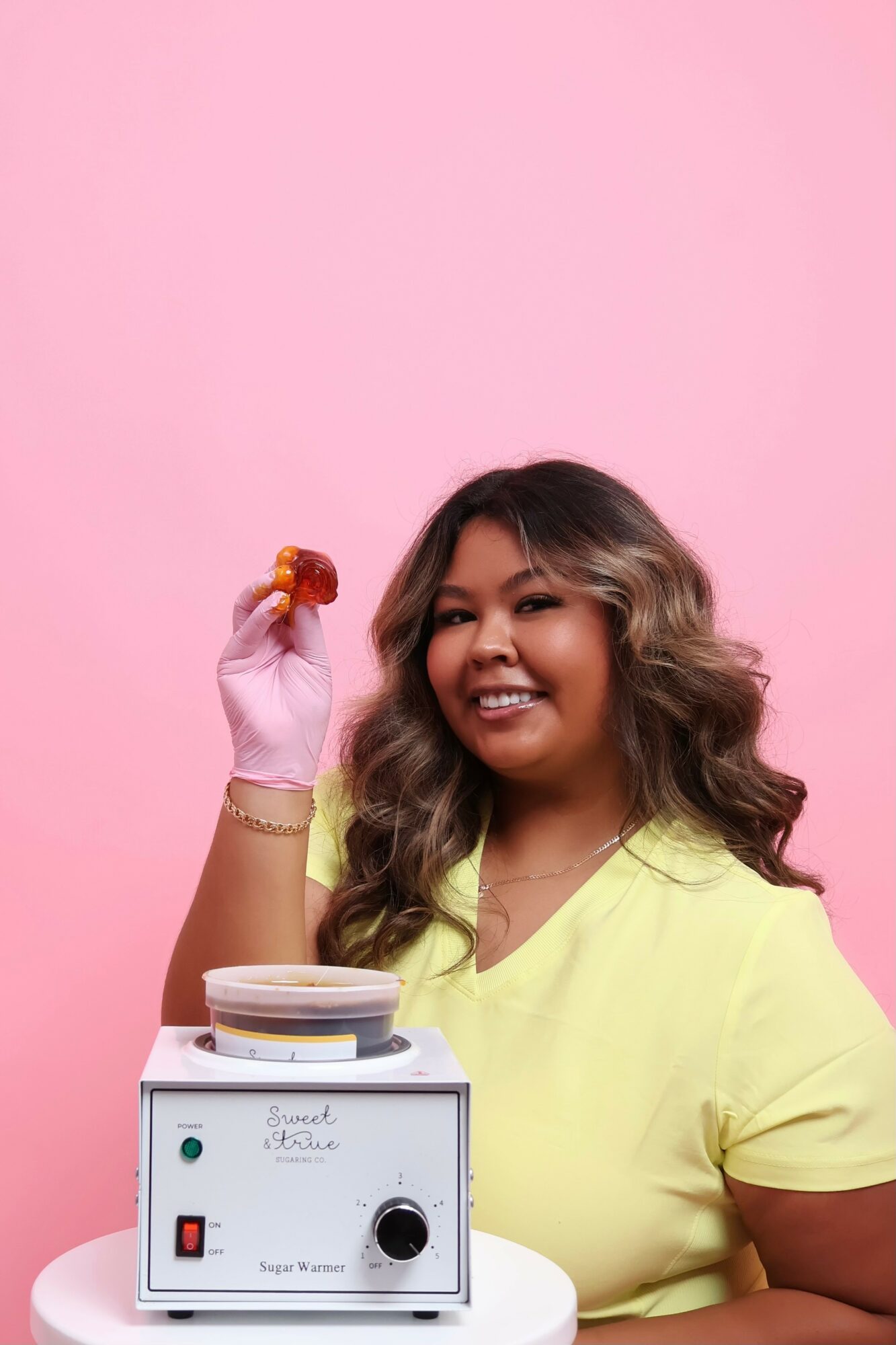 Smiling woman with wavy hair holding a small object, standing next to a machine against a pink background.