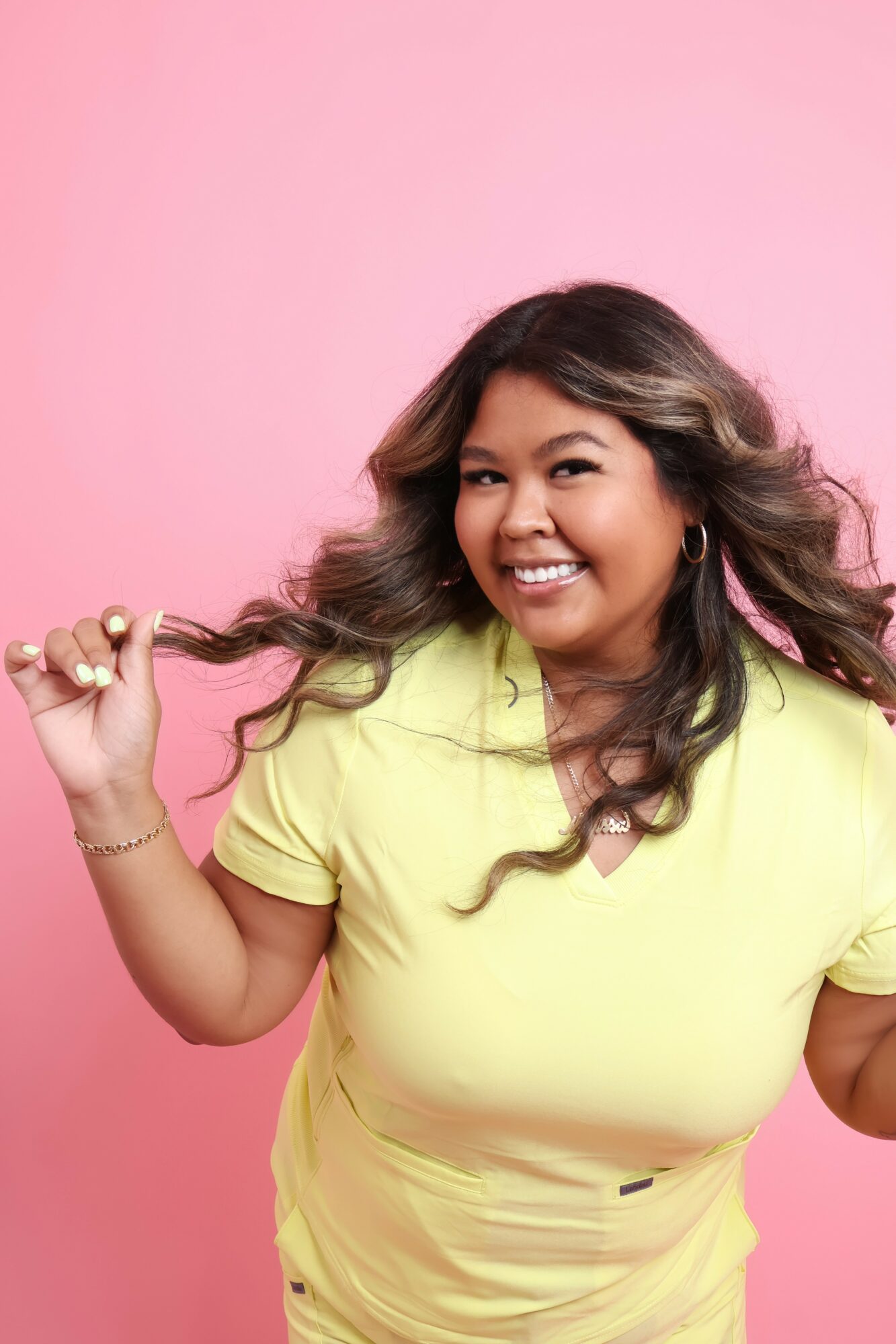 Smiling woman with long wavy hair wearing a yellow top, holding a strand of her hair, against pink background.