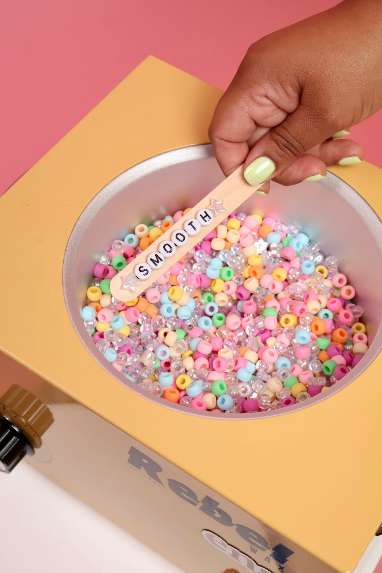 Hand holding a stick labeled 'SMOOTH' over a bowl of colorful beads, with a pink background and yellow box.