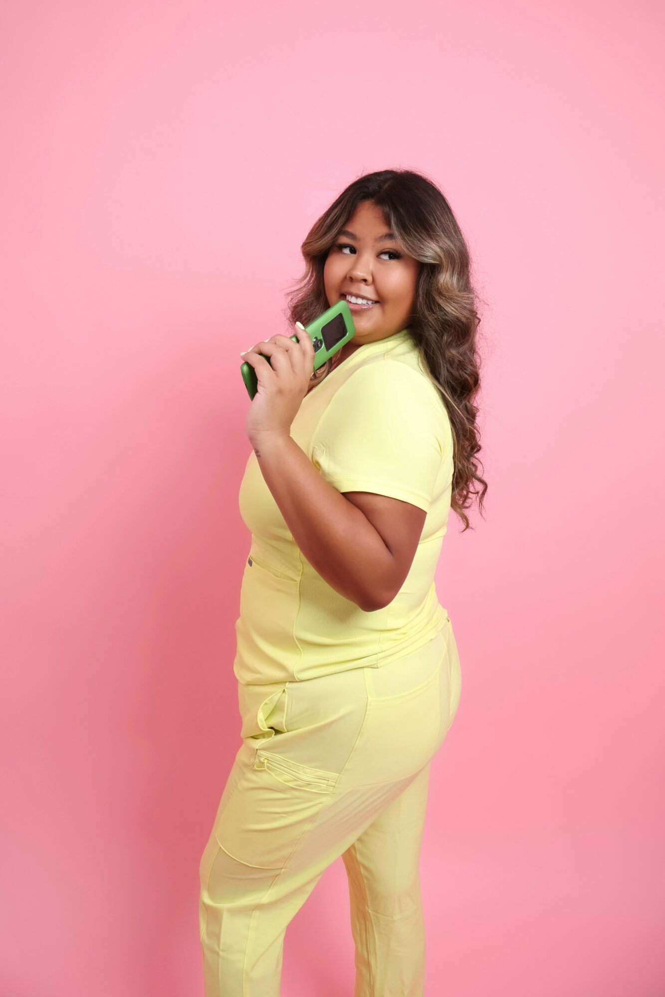 Smiling woman in yellow dress holding a green phone against pink background.