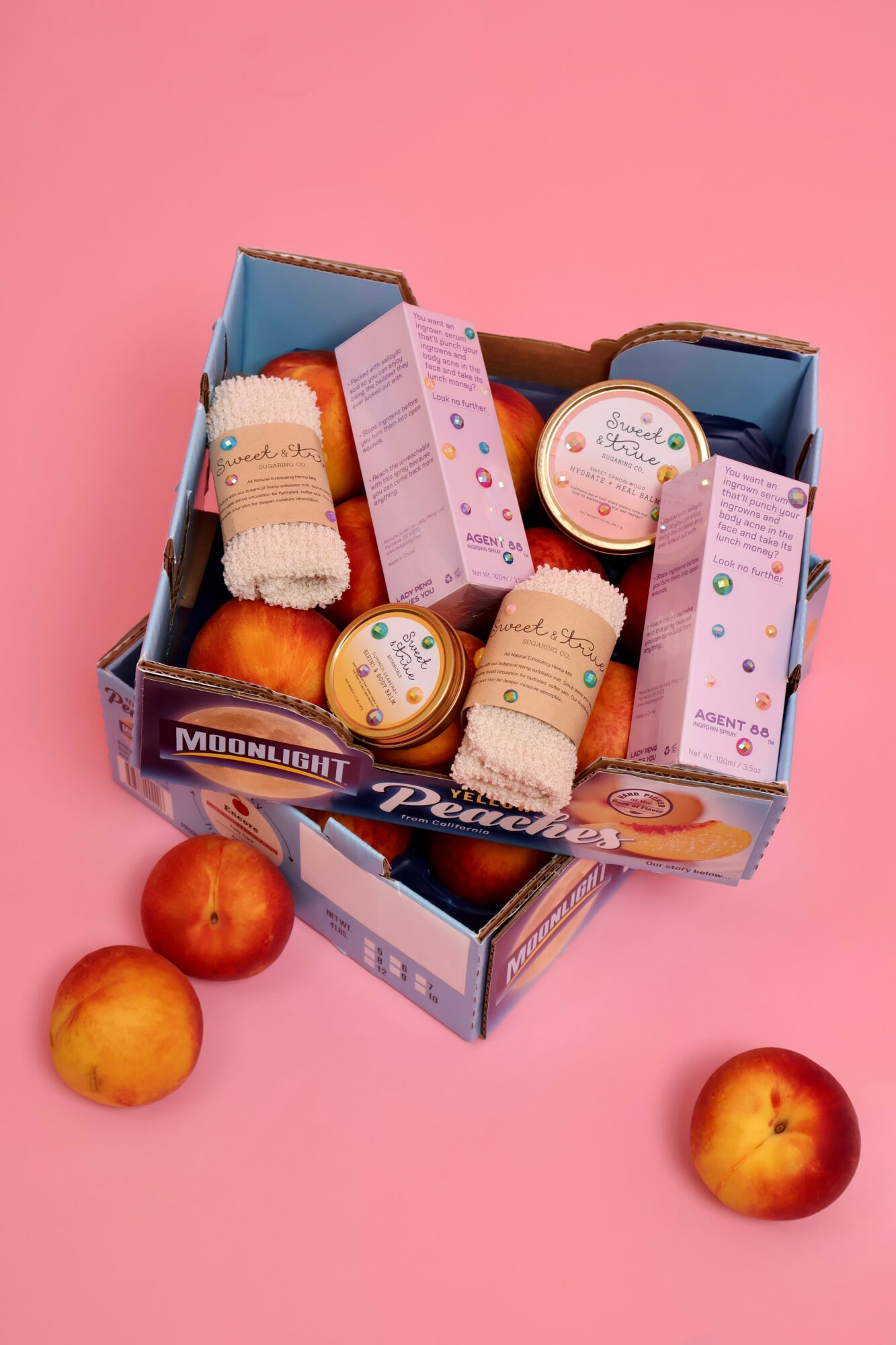 Open box of skincare products with three peaches on pink background.