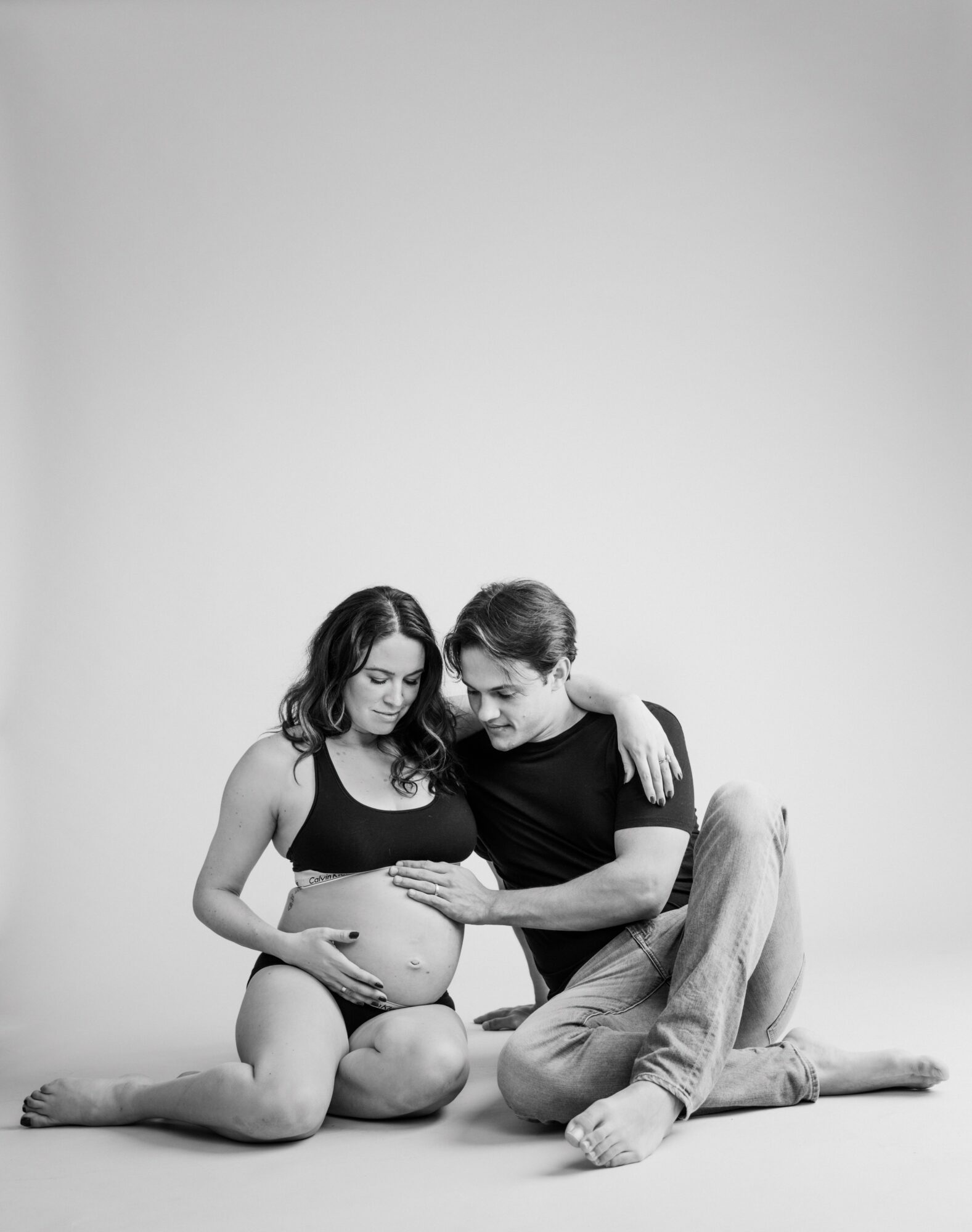 A couple sitting on the floor, with the woman pregnant, touching her belly, and the man leaning in, touching her belly.