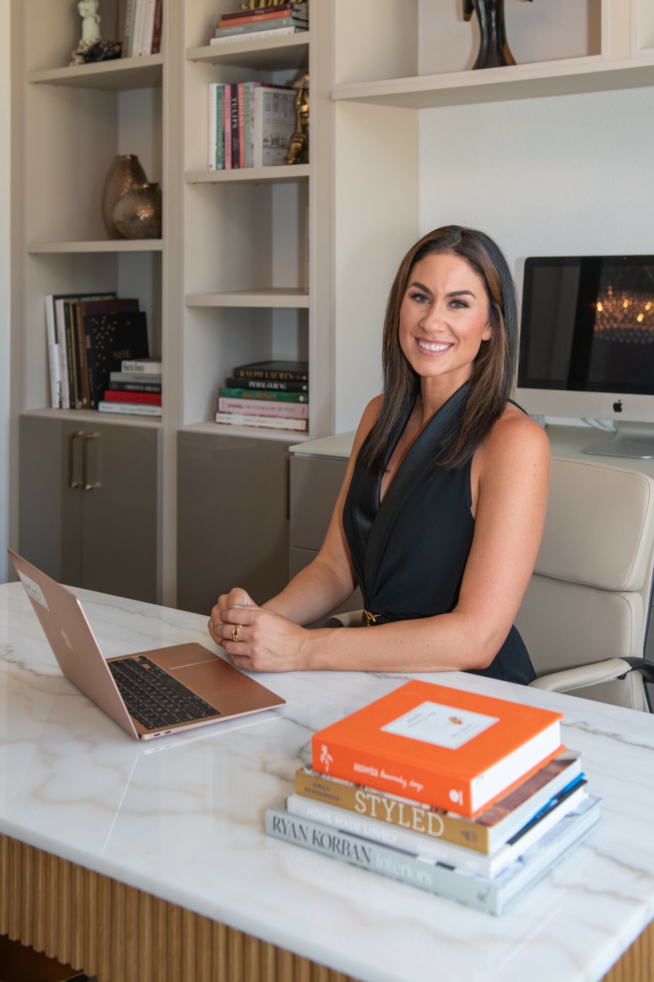 Woman sitting at a desk with a laptop and stacked books, smiling, in a room with bookshelves in the background.