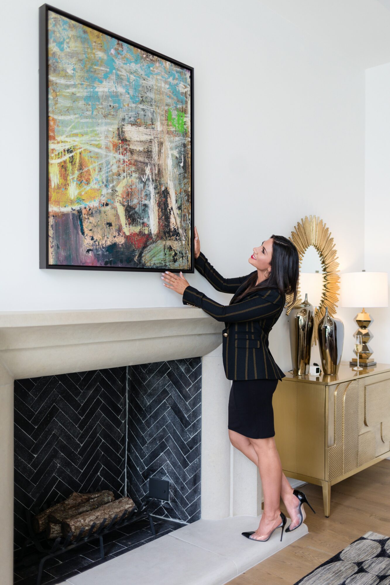 Woman in black blazer and skirt adjusts large colorful abstract painting above fireplace in a modern living room.