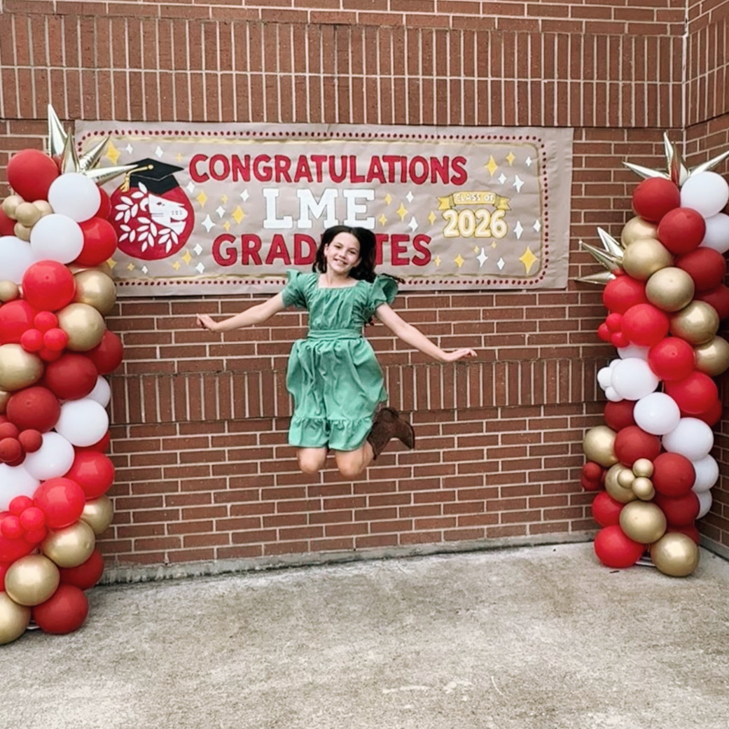 Young girl jumping in front of a graduation celebration banner with balloon decorations on brick wall, smiling, arms outstretched.