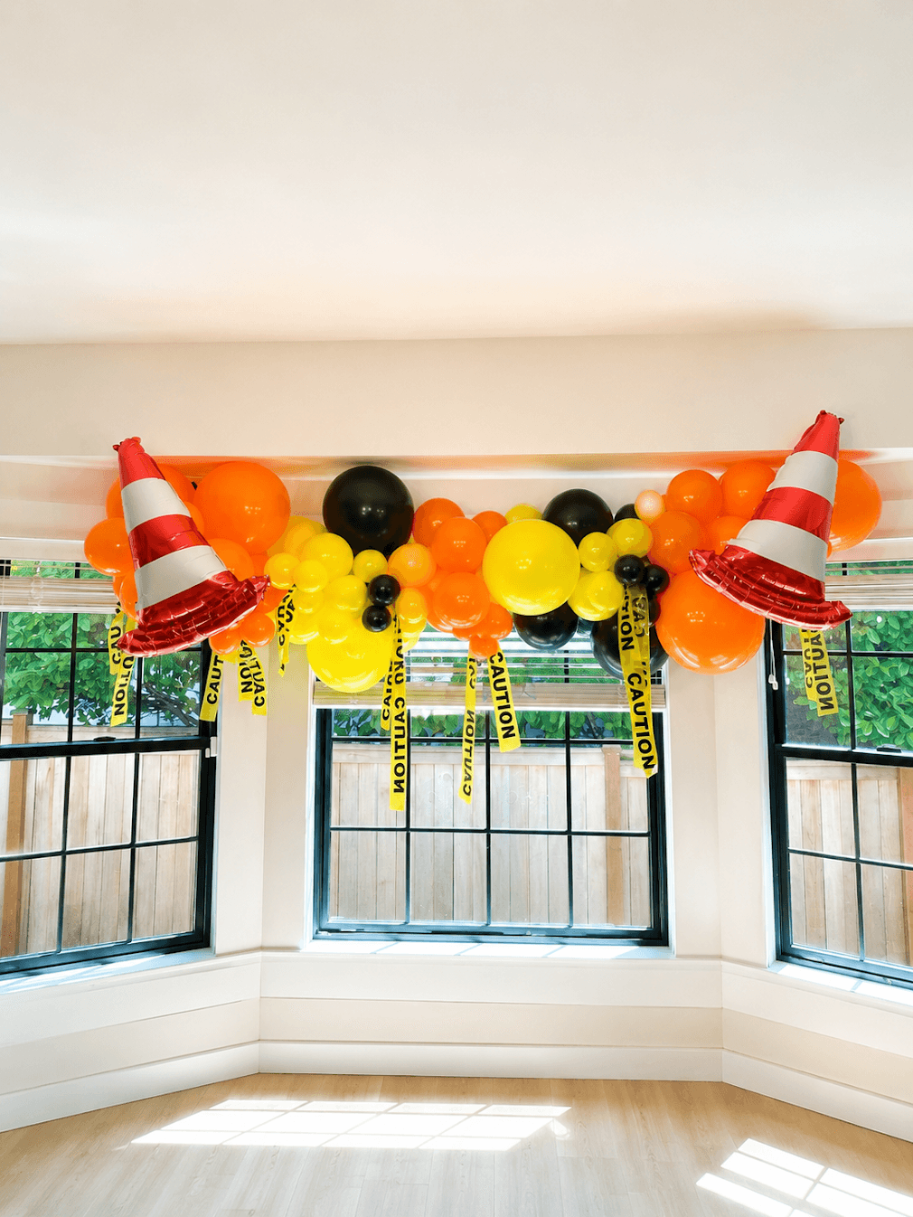 Decorative balloon arrangement with traffic cones and caution tape in a room with three windows, sunlight, and a white ceiling.