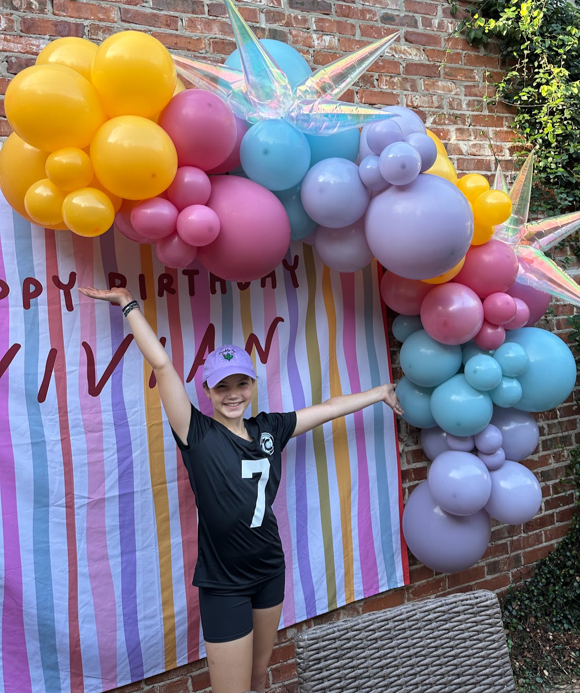 Child in sports uniform smiling with arms outstretched in front of colorful balloon arch and striped backdrop.