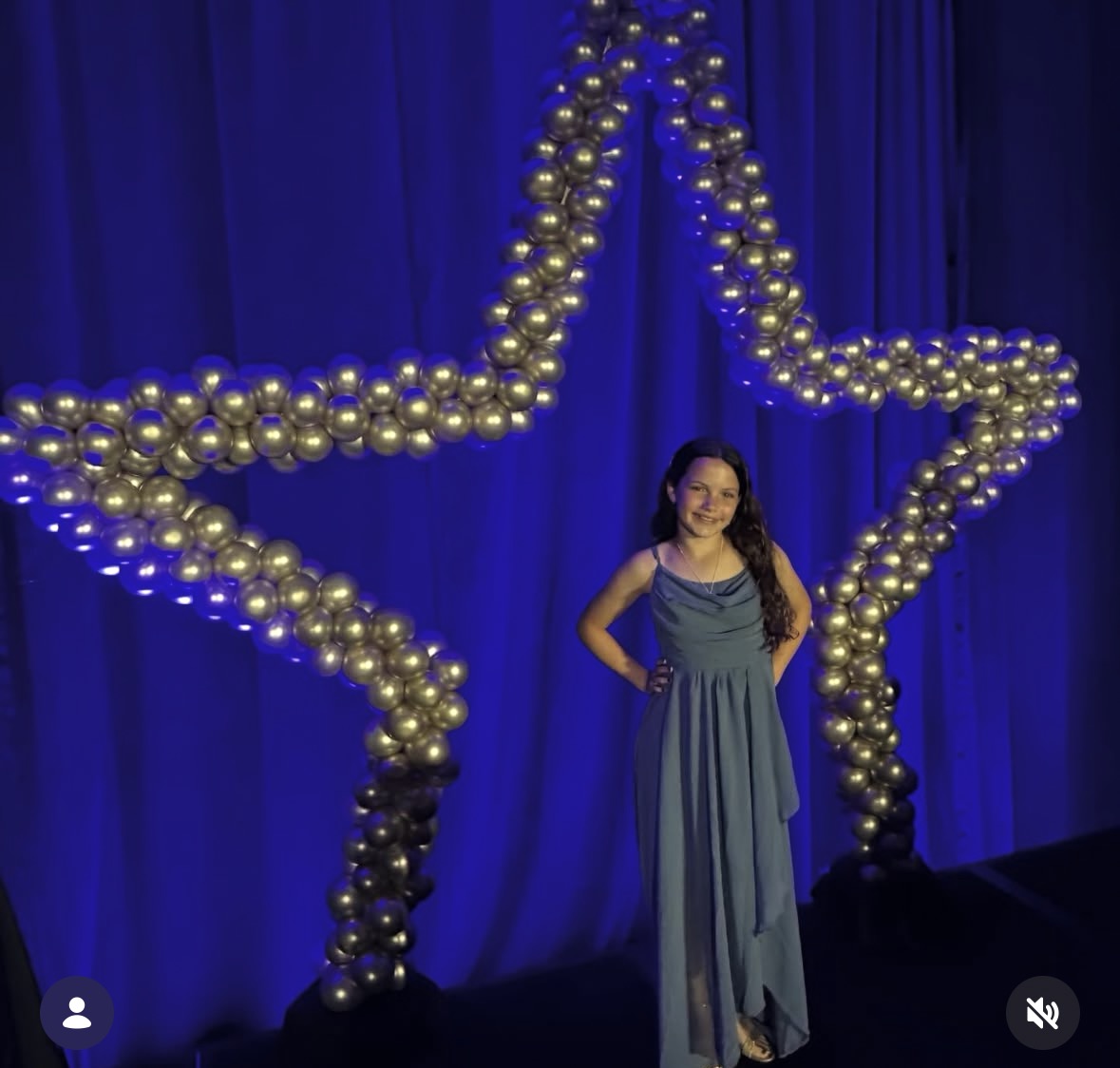 Girl in a blue dress standing in front of a large star made of gold balloons against a blue curtain backdrop.
