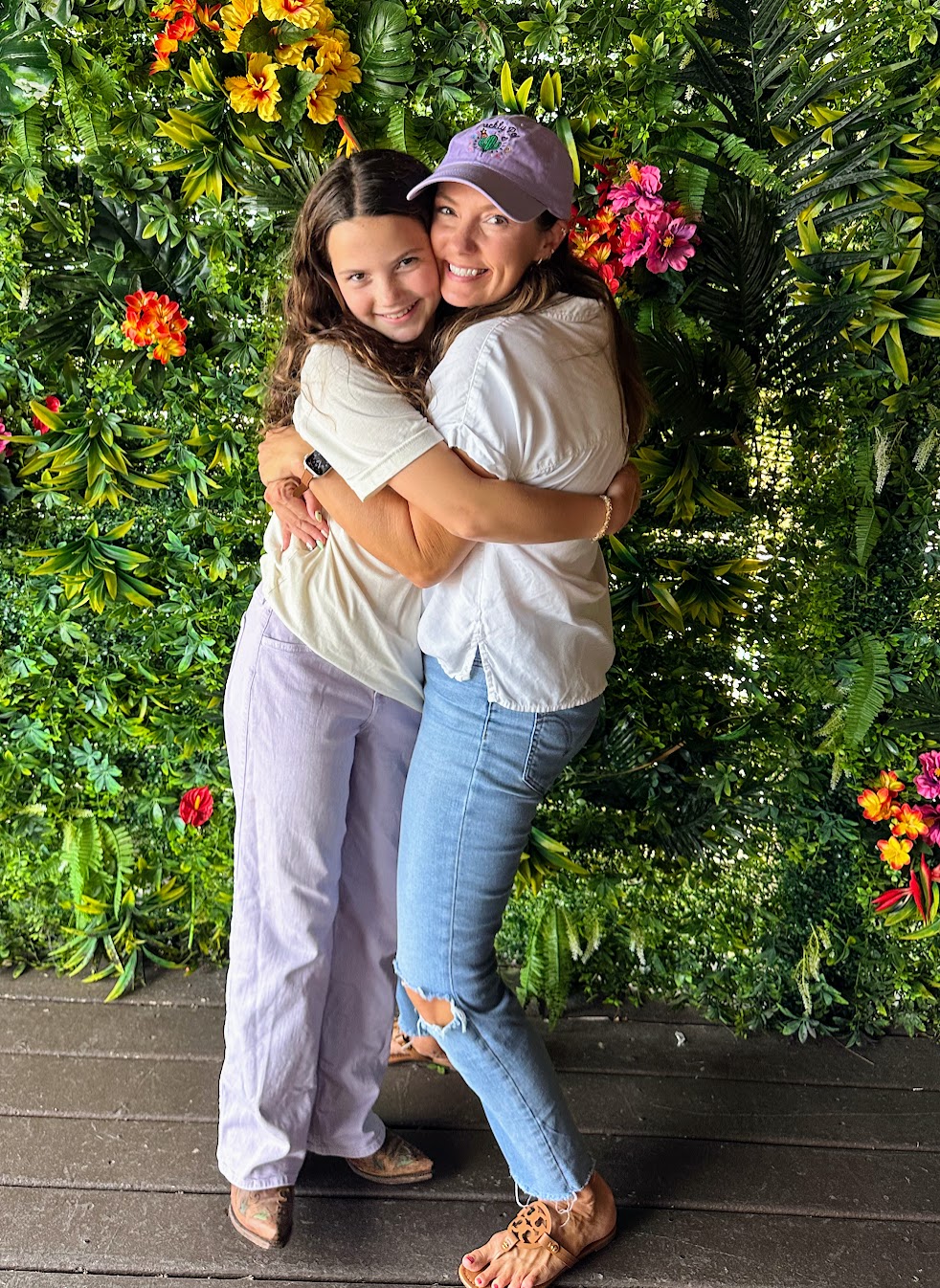 Two women hugging and smiling, standing in front of a lush green floral background with colorful flowers.