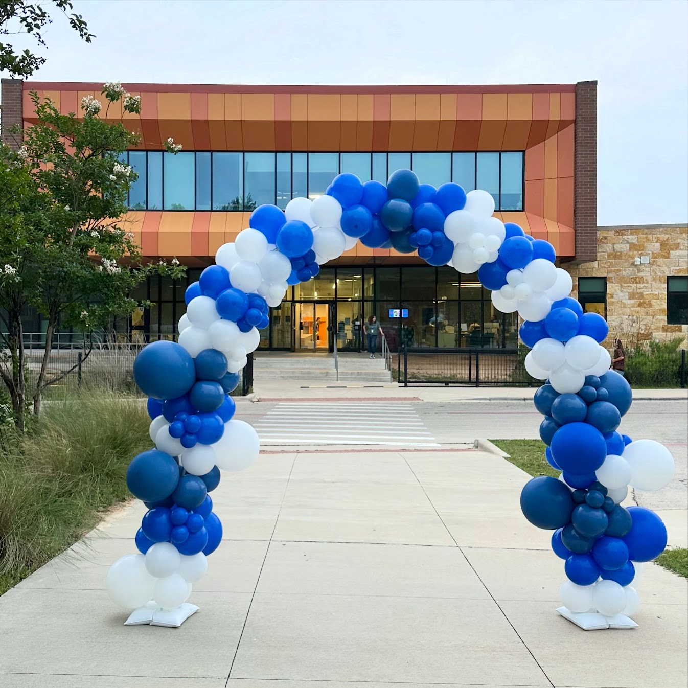 Balloon arch with blue and white balloons in front of a modern building entrance.