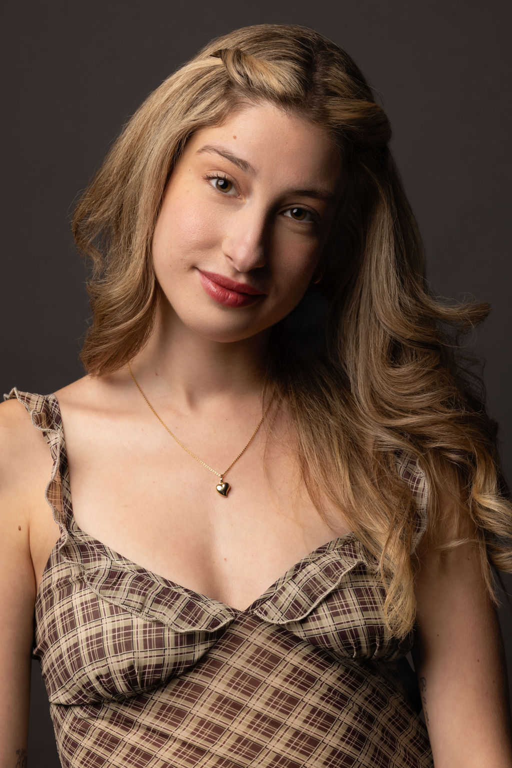 Young woman with long wavy hair wearing a plaid top and necklace, smiling softly against a dark background.