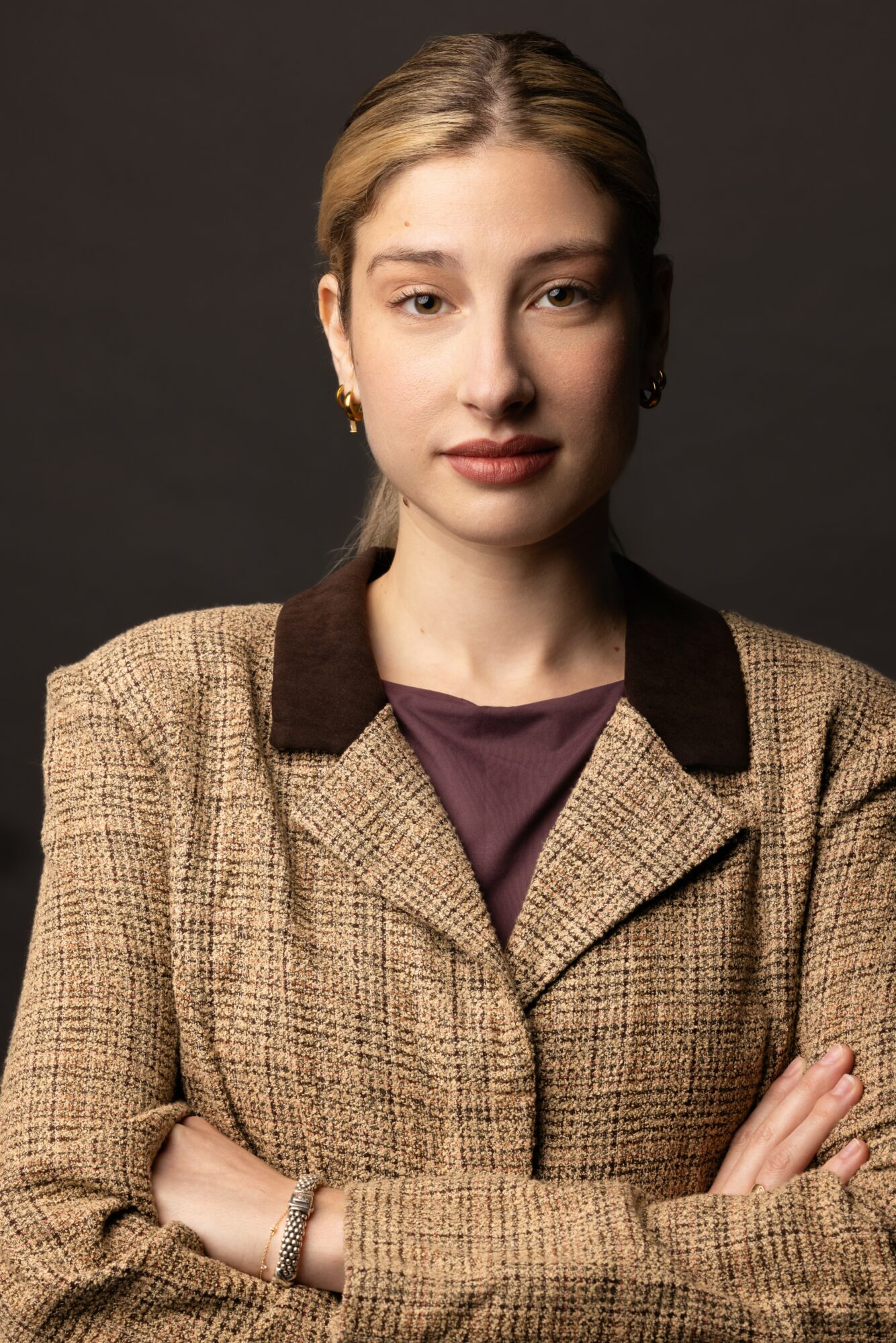 Young woman with blonde hair in a bun, wearing a brown checkered blazer with dark collar, crossing arms, neutral expression