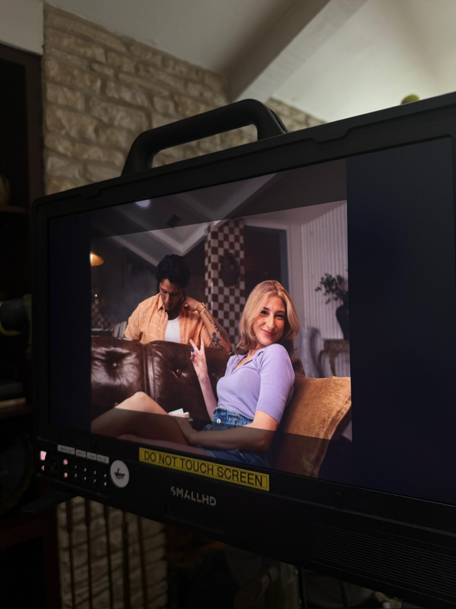 Person with blonde hair smiling, sitting on a couch, with another person in the background, in a cozy room.