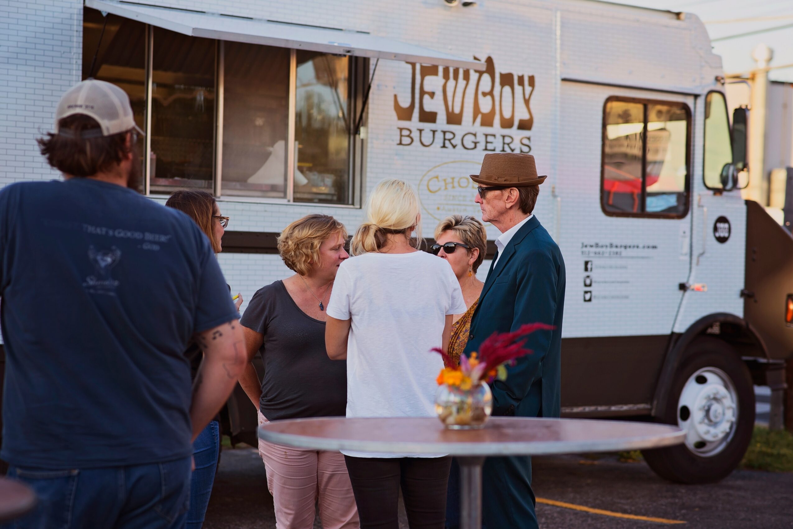 Group of people talking near a food truck with a sign that reads 'Jewboy Burgers', outdoor setting, table with a flower arrangement.