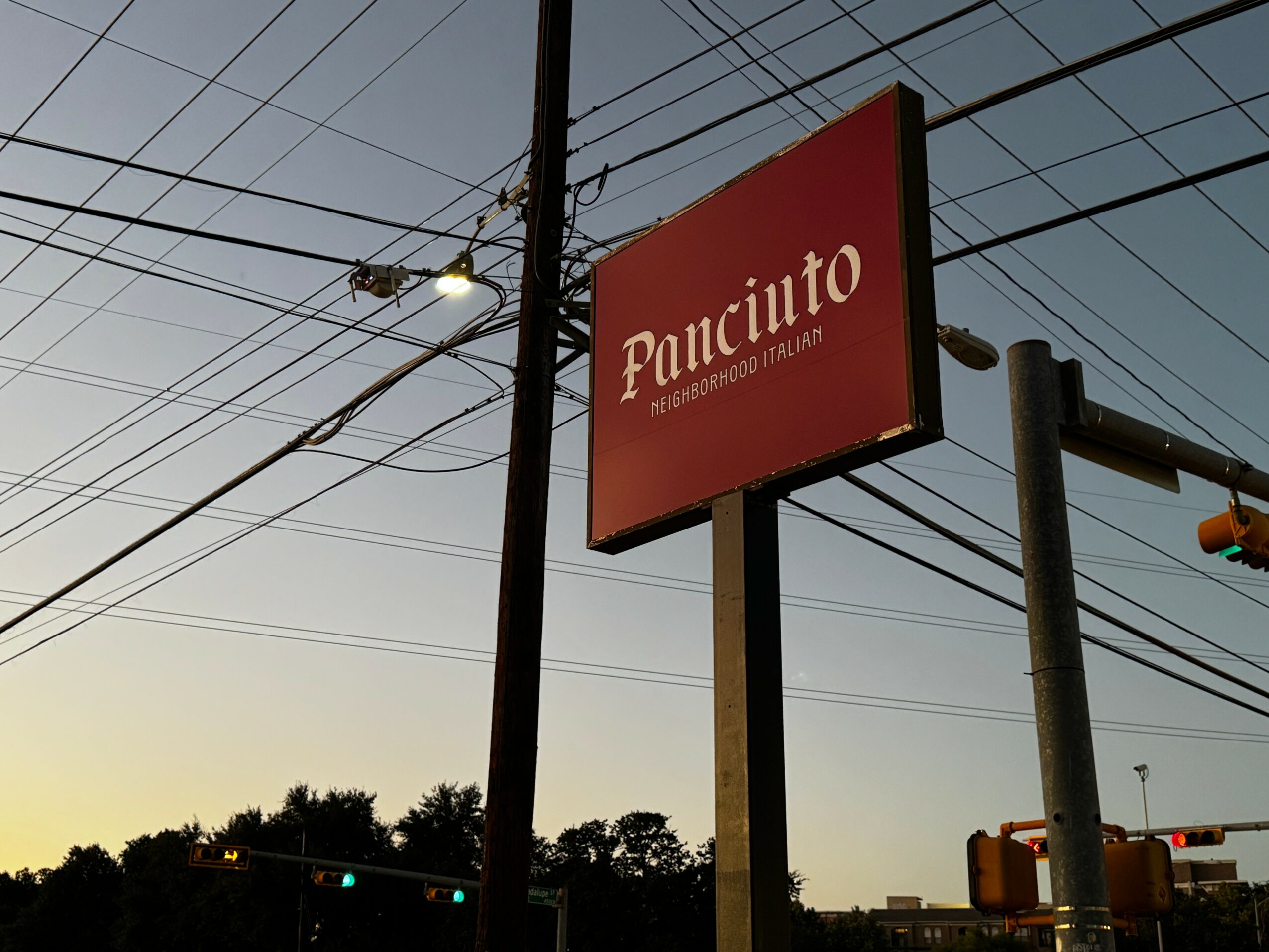 Red sign with white text reading 'Pancito' hangs above a street, with power lines and a traffic light in the background.