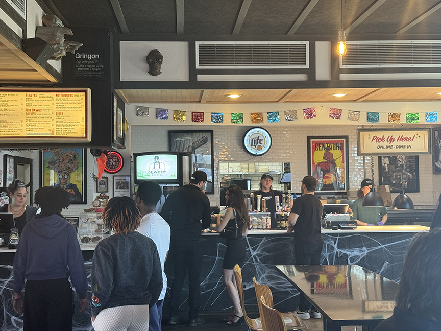 People standing at a bar counter inside a cafe or restaurant, with colorful posters and a menu board behind the counter.