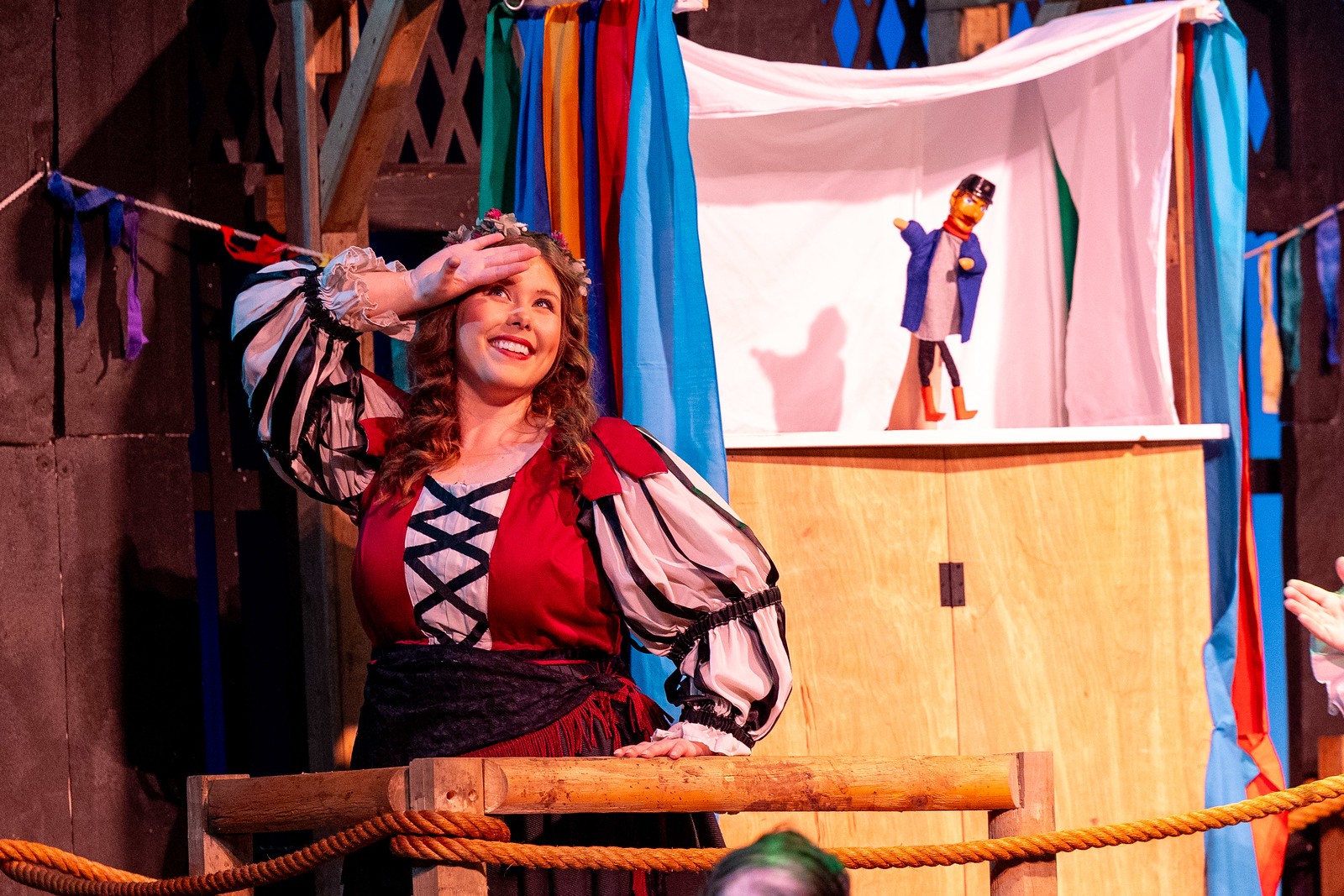 A woman with long hair in a colorful costume smiles on stage with a puppet show in the background.