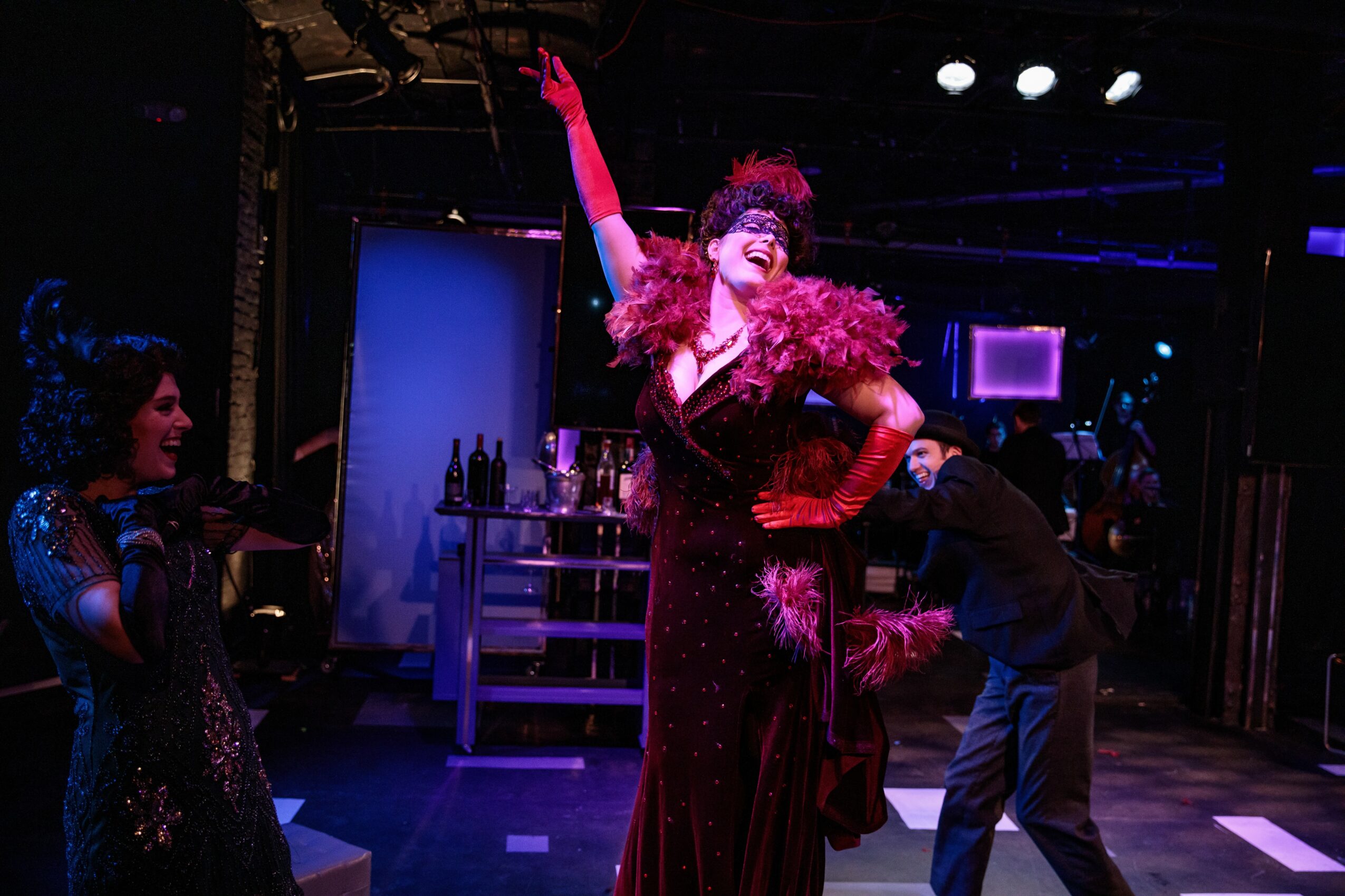 Performer in a glamorous costume with feathers and gloves on stage, with a person in dark clothing nearby.