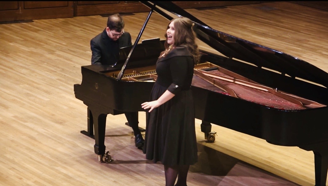 A woman singing and a man playing piano on stage with wooden flooring.