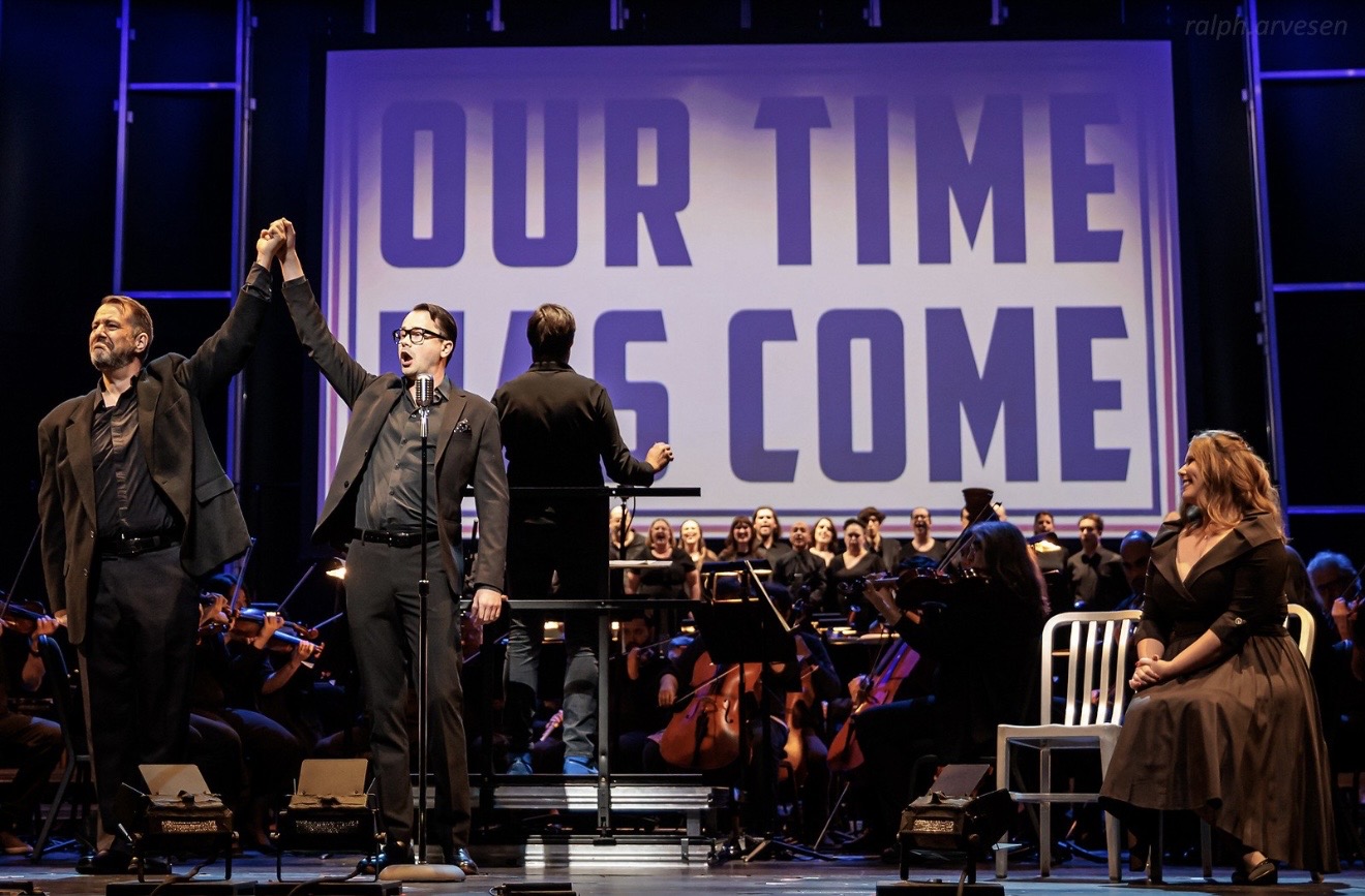 Three people on stage with an orchestra and a large screen displaying 'OUR TIME HAS COME'.