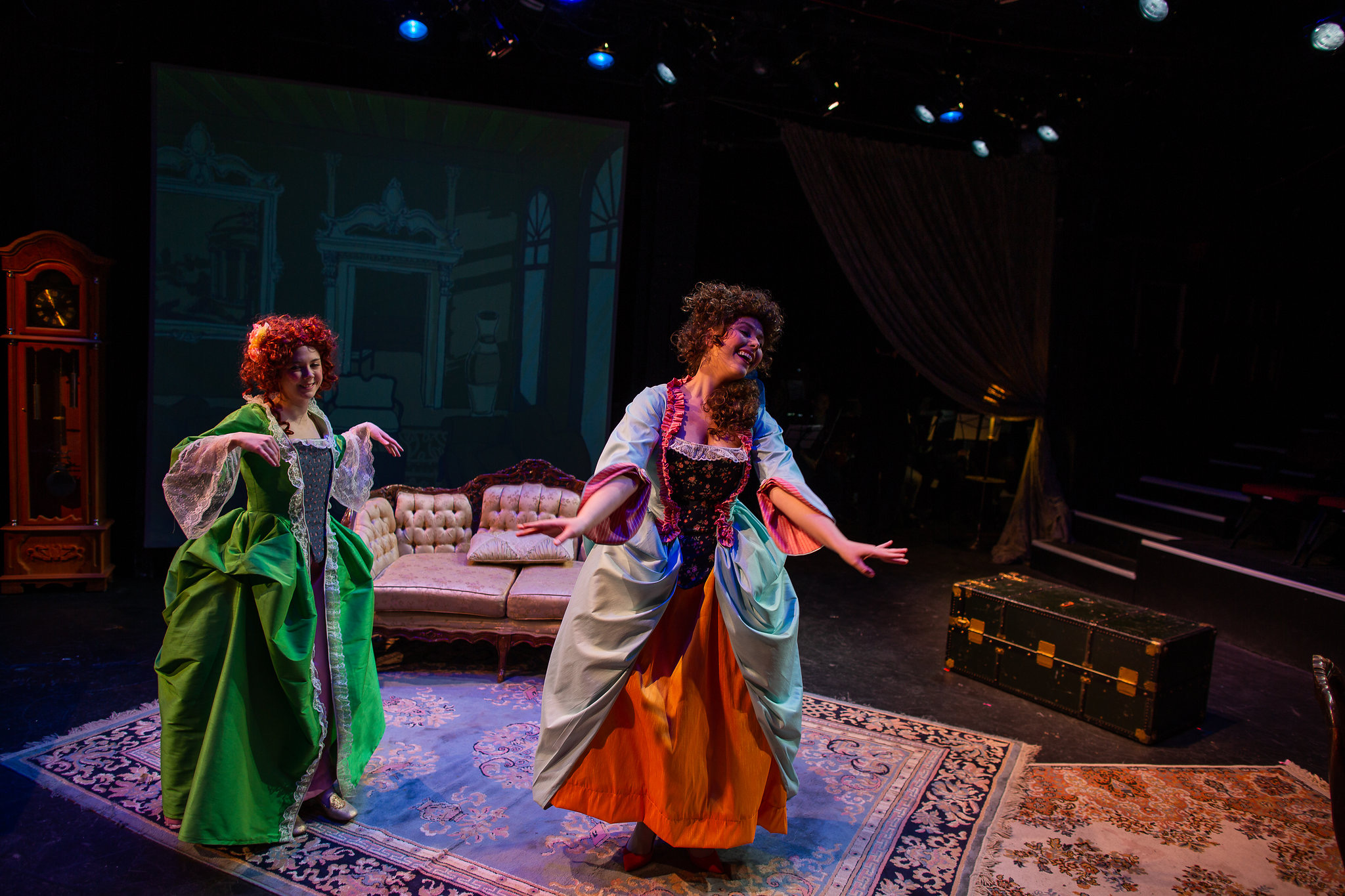 Two actors in colorful costumes perform on stage with a sofa and a screen in the background.