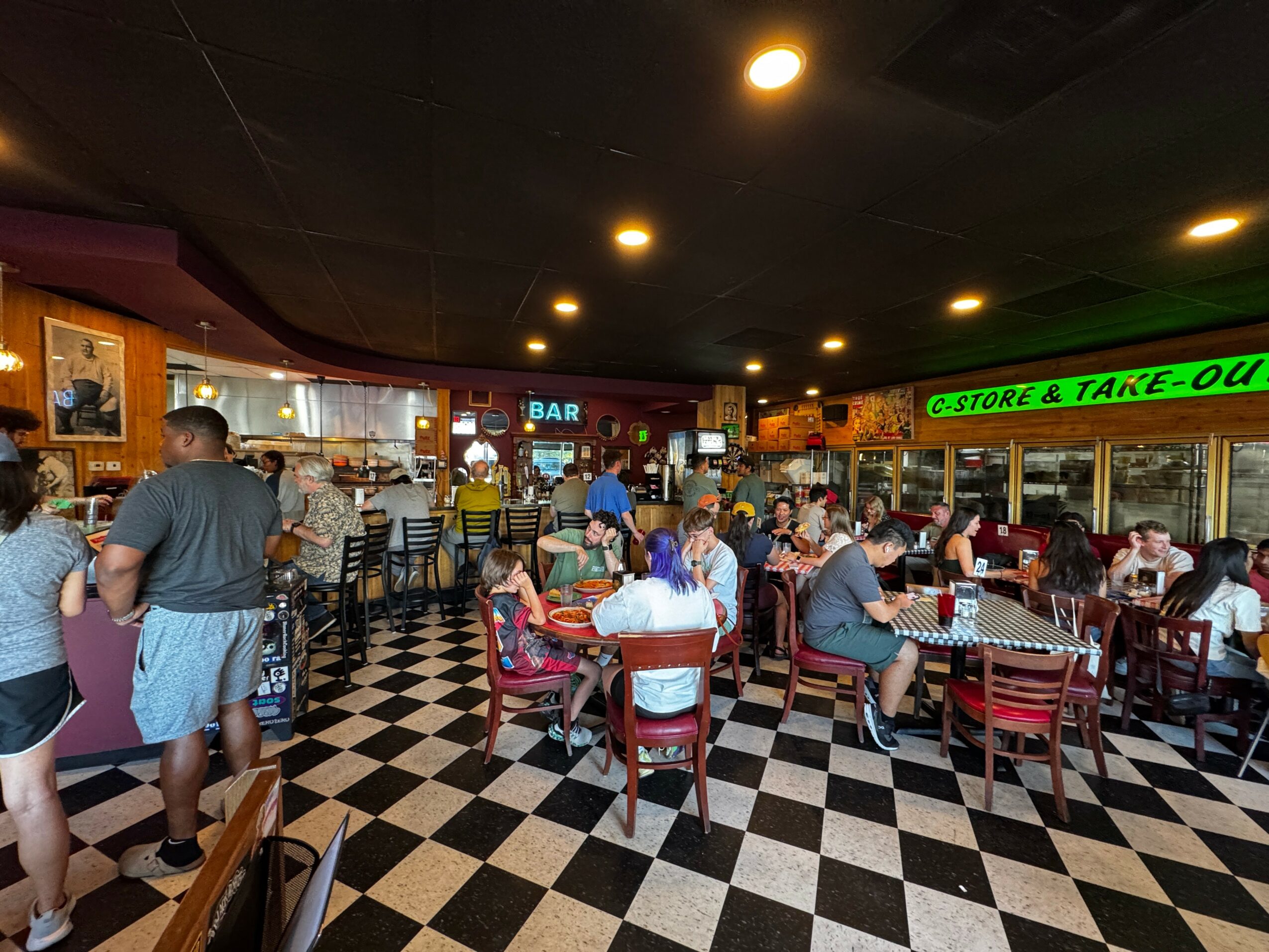 People dining at tables in a restaurant with checkered floor, bar area, and bright signs, some standing and some seated.