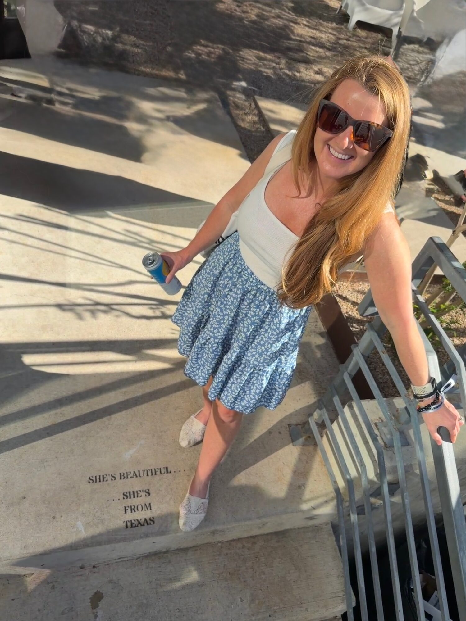 A woman wearing sunglasses and a blue patterned skirt stands beside a metal railing on a sunlit walkway.