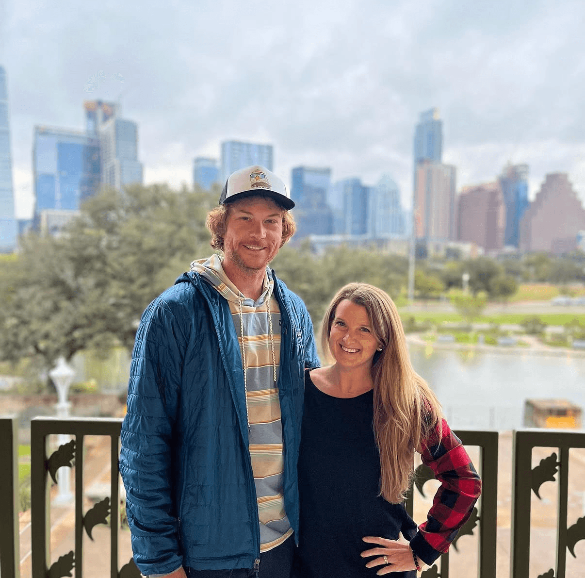 Two people stand by a railing, with a lake, trees, and city skyline behind them.