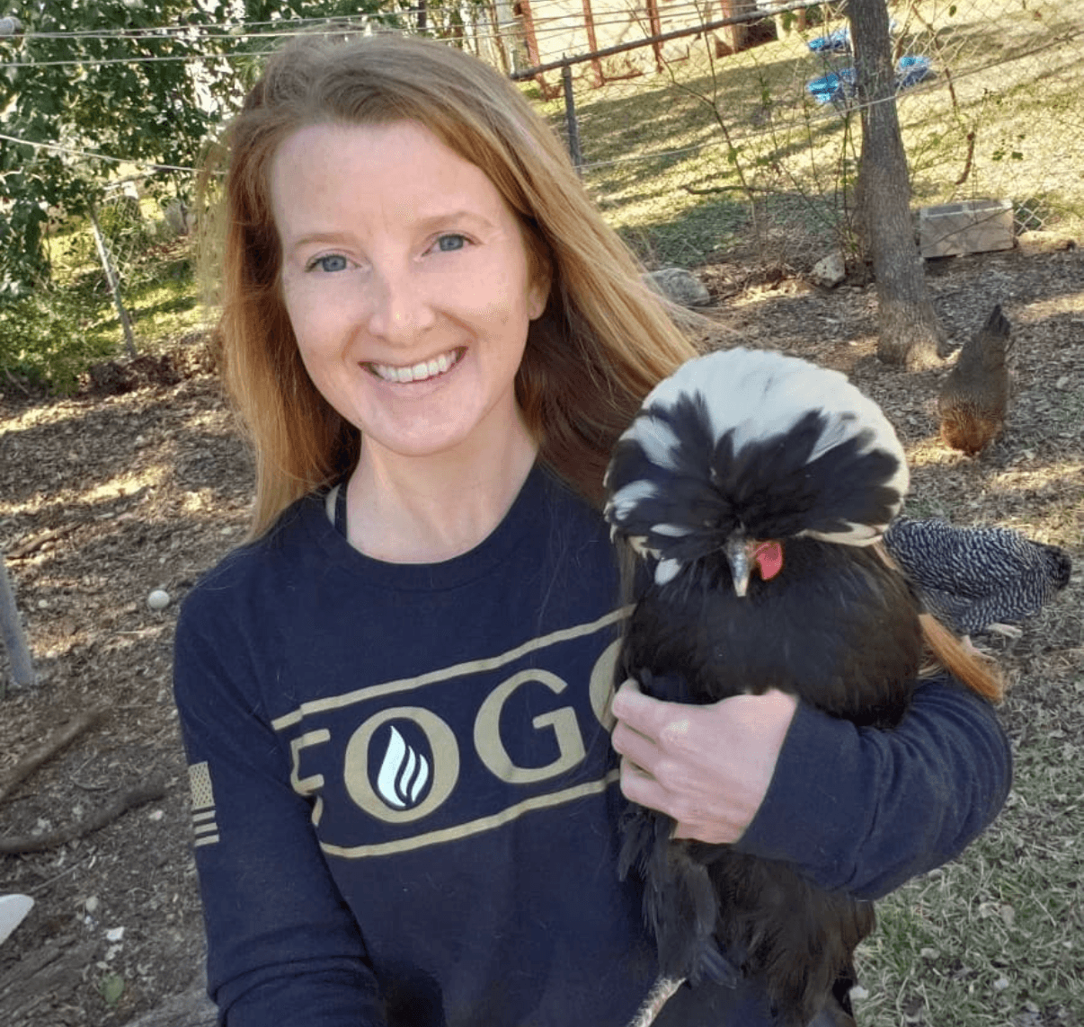 A person holds a black-and-white crested chicken outdoors, with other chickens and a fenced yard behind them.