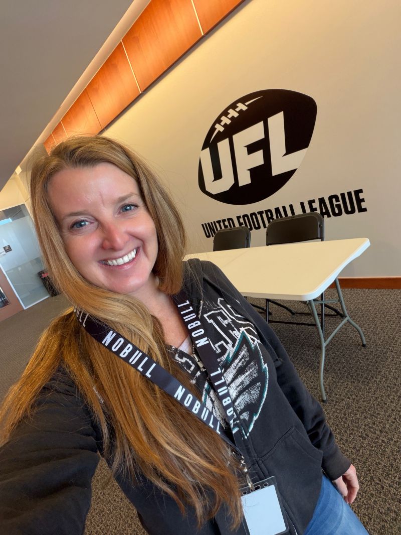 Smiling person wearing a lanyard poses beside a wall logo and classroom tables.