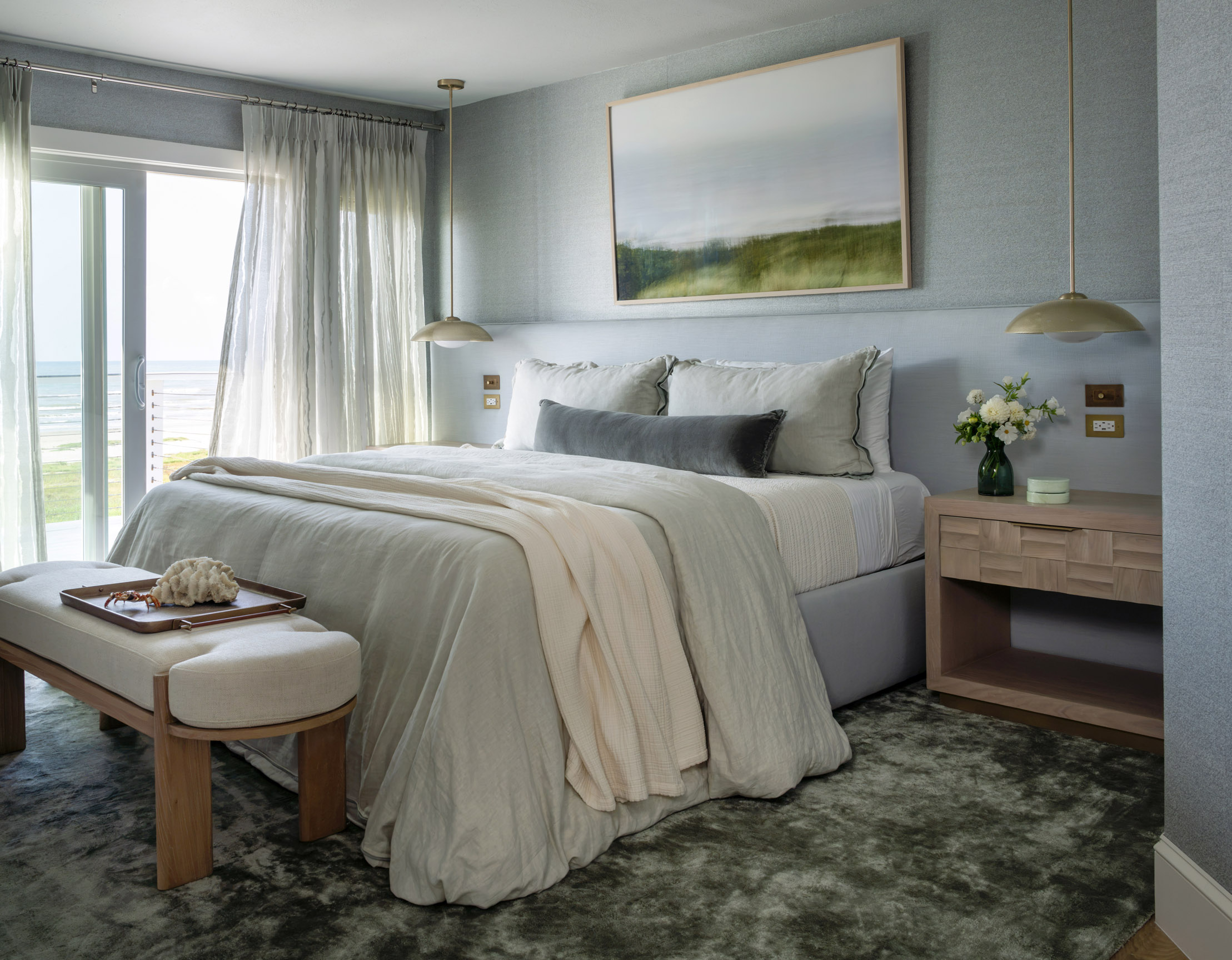 A bedroom with a large bed, artwork above, bedside table with flowers, and a window with curtains.