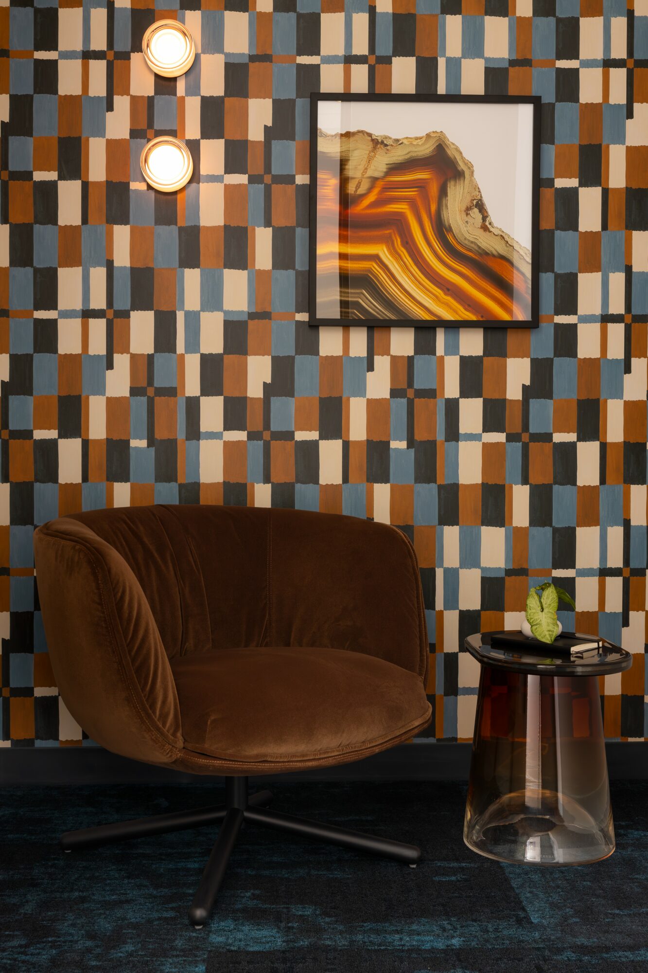 Brown armchair with a small round table holding a plant, against a patterned wall with a framed artwork.
