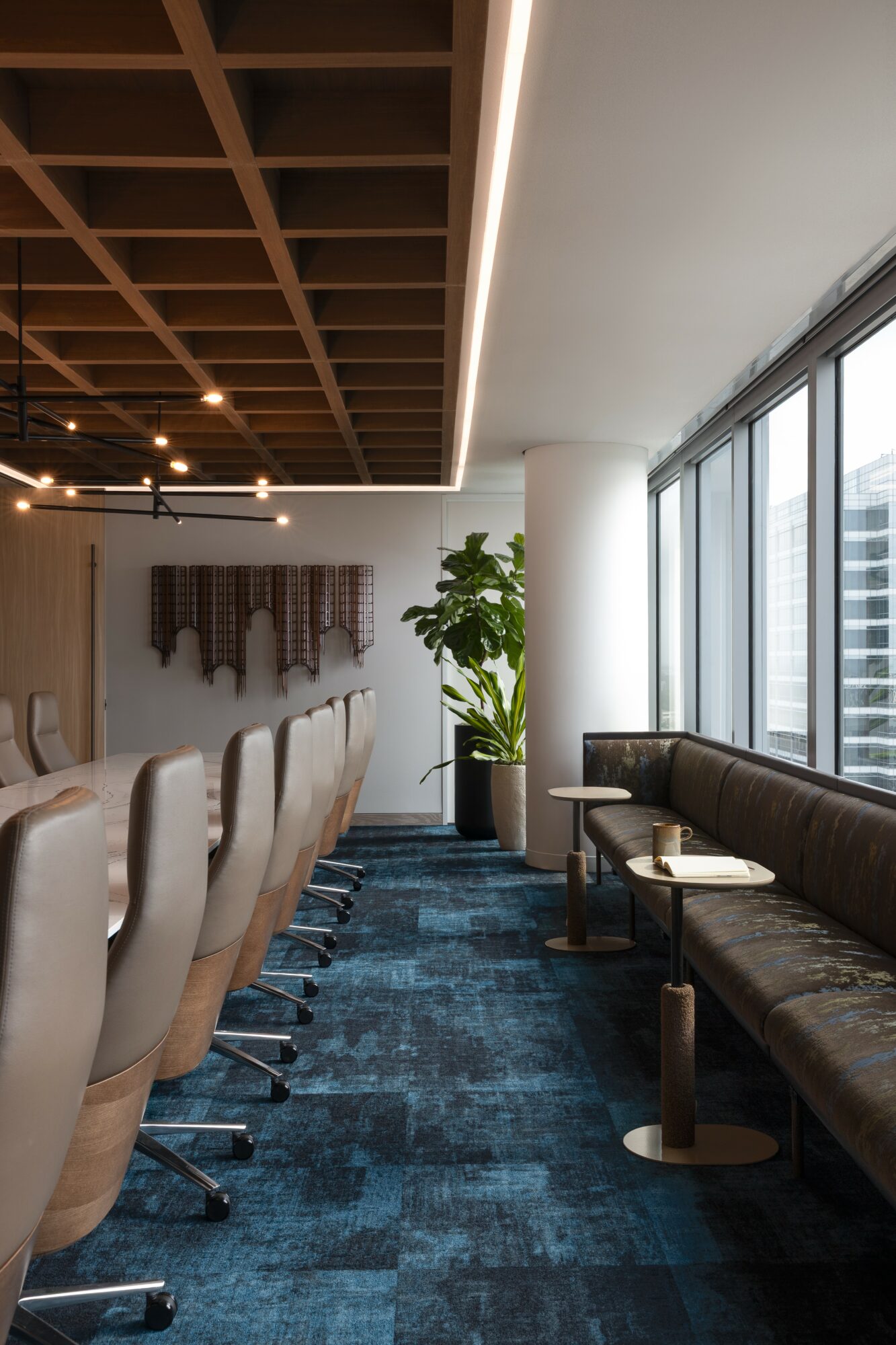 Interior of a modern office or lounge with chairs, a bench, large windows, and a potted plant, viewed from the side.