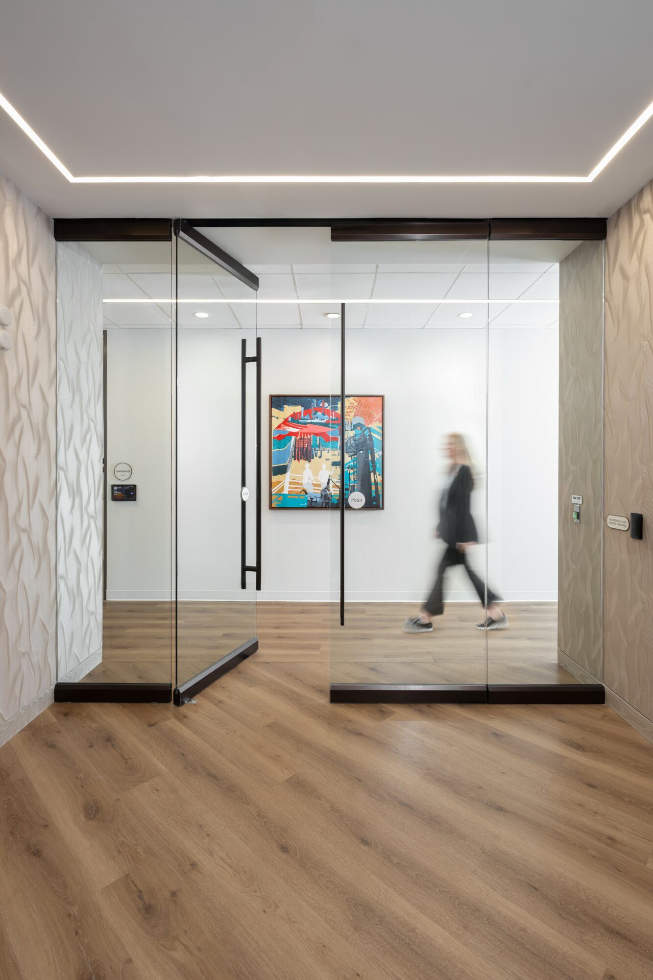 Person walking past glass doors in a modern interior with artwork on the wall, wooden floor, and ceiling lighting.