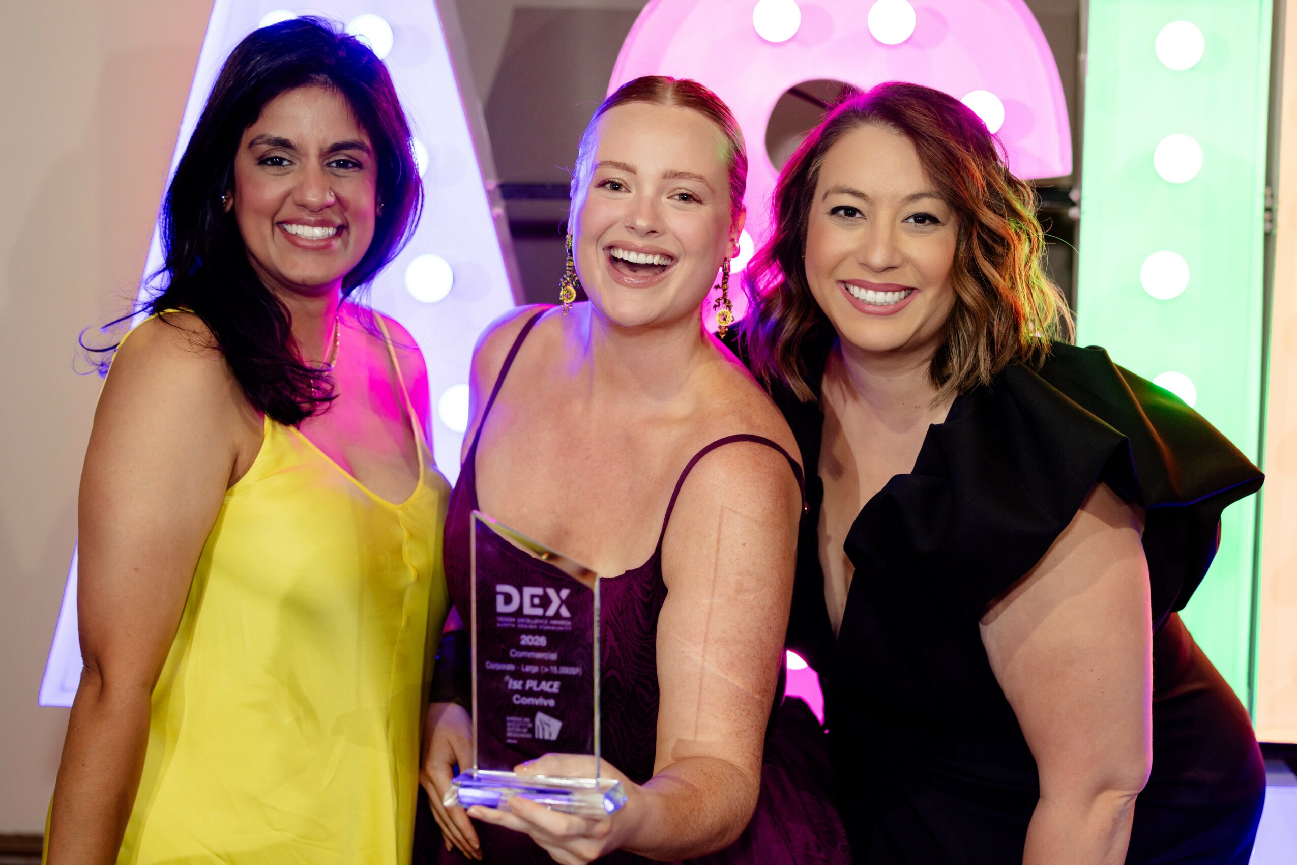 Three women smiling, standing close together, with colorful background lights and decorations.