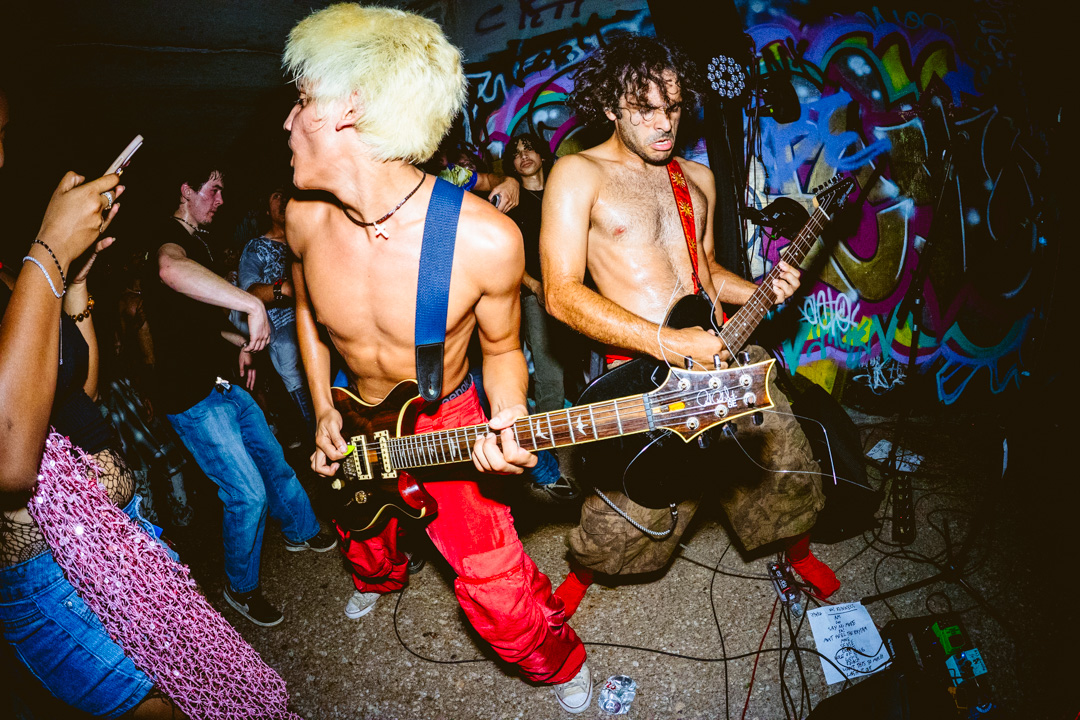 Two shirtless musicians perform with guitars in front of a graffiti-covered wall, surrounded by an audience. Colorful lighting highlights the scene.