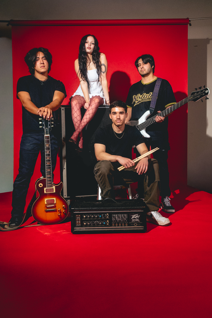 Four people with musical instruments pose against a red backdrop, one sitting on a box, others standing, with a guitar and drumsticks.