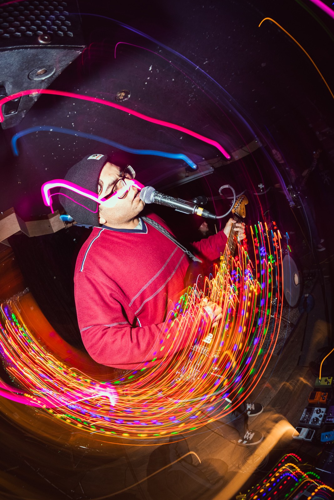 Person wearing glasses and a red jacket, holding a microphone, surrounded by colorful light trails, performing on stage.
